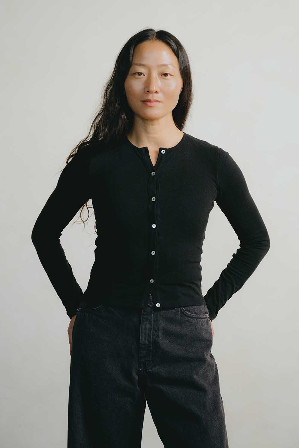 A woman modeling a black long-sleeve button-front top with dark wide-leg jeans, standing against a plain light gray background.