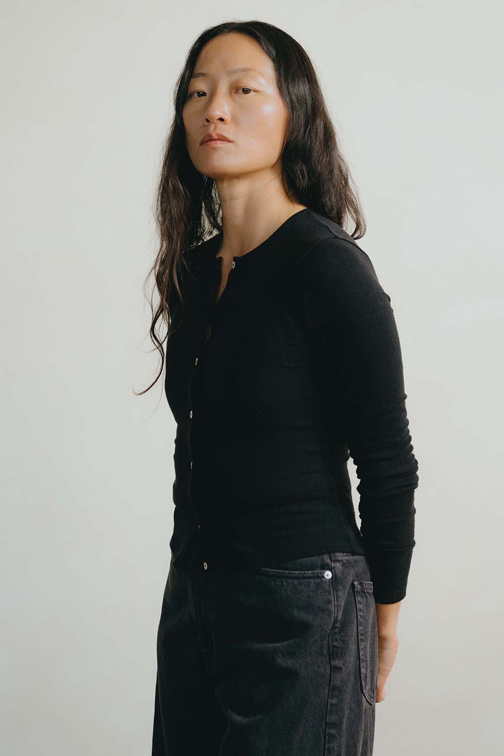 A woman with long dark hair is shown in profile wearing a black long-sleeve button-front top and dark jeans against a plain light gray background.