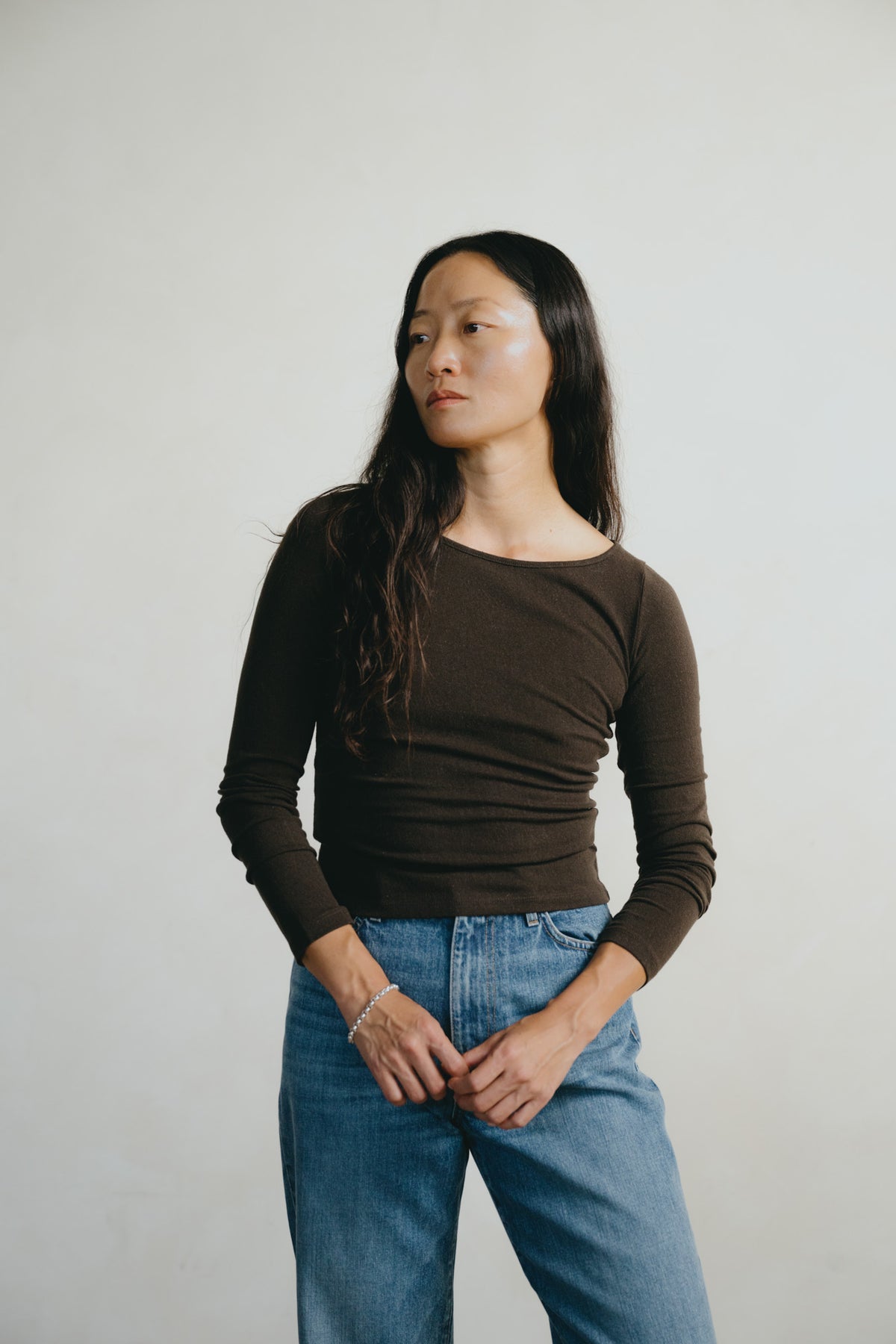 A woman with long dark hair wearing a dark brown long-sleeve crewneck top and light blue jeans, standing against a plain light gray background.