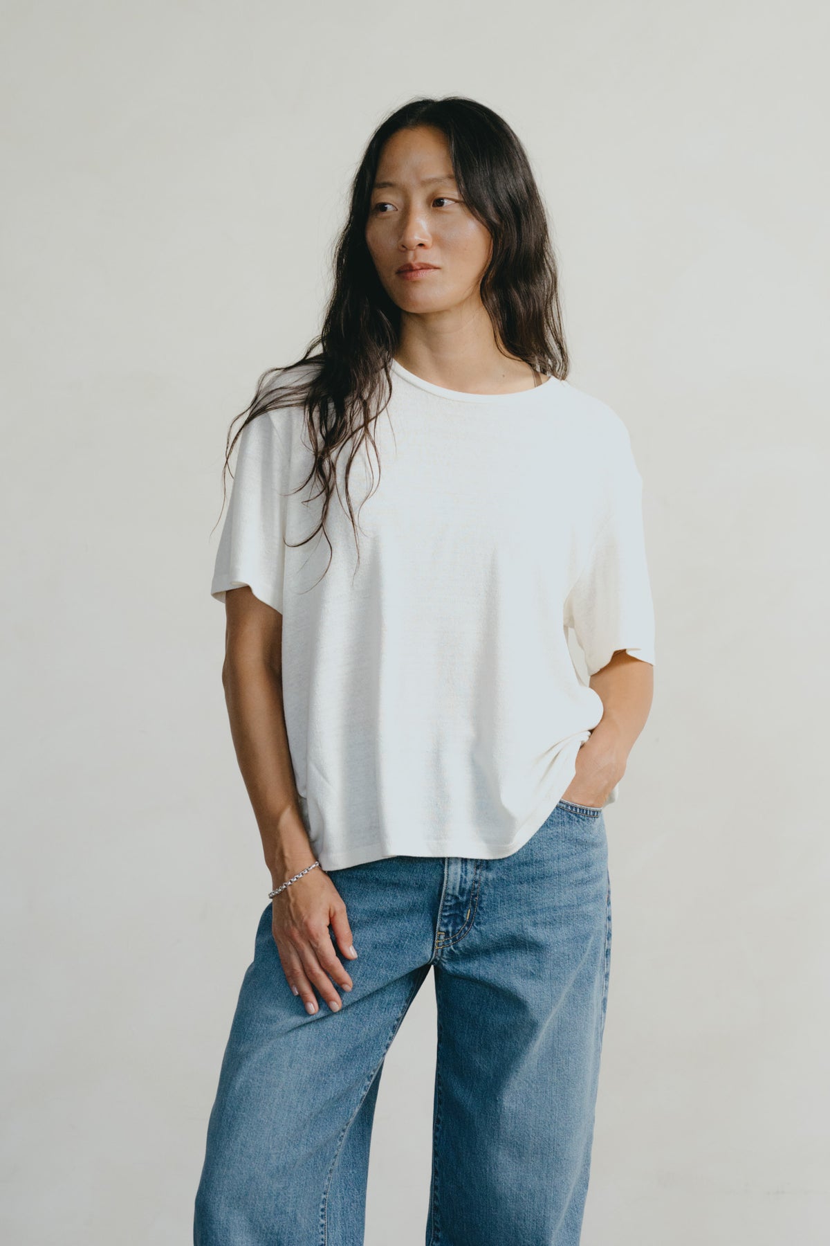 A woman modeling a loose-fitting white short-sleeve t-shirt paired with blue jeans, standing against a plain light gray studio background.