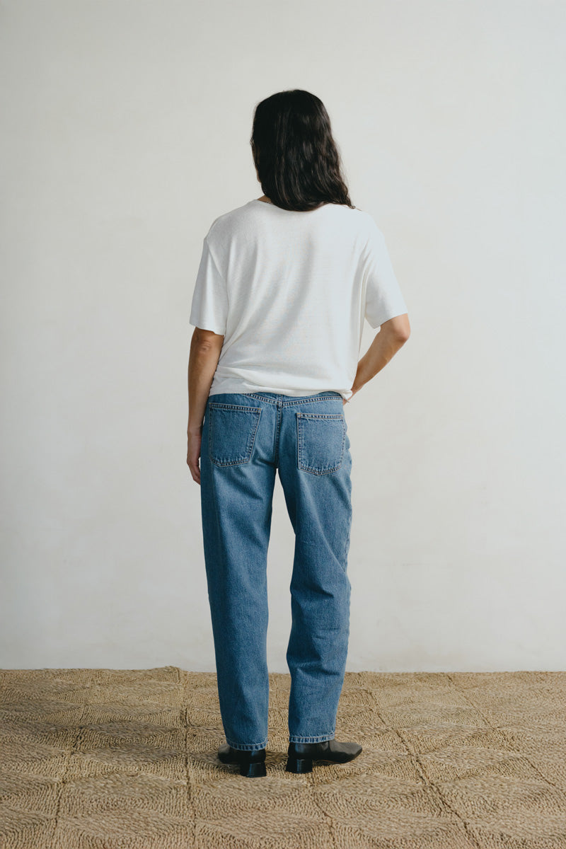 A person standing with their back to the camera, wearing a white short-sleeve t-shirt and blue straight-leg jeans, standing on a textured beige floor against a plain off-white wall.