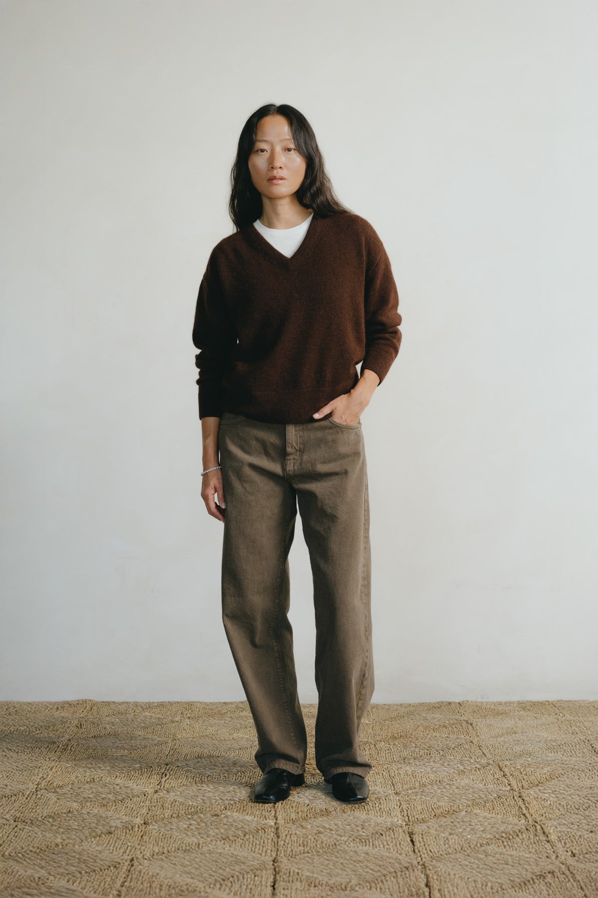 A woman stands in a studio wearing a dark brown V-neck sweater over a white undershirt, paired with brown wide-leg trousers and black shoes, against a plain light gray wall and woven jute floor.