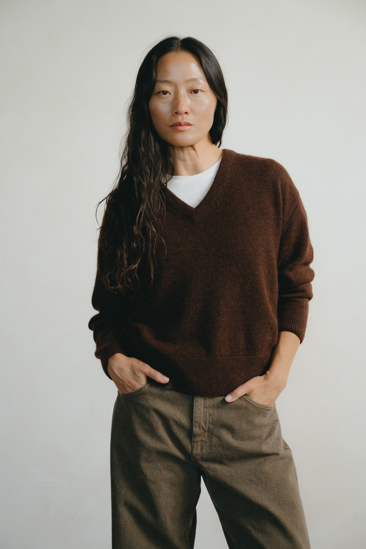 A woman with long dark hair stands against a plain light gray background wearing a dark brown v-neck knit sweater over a white t-shirt and brown pants.