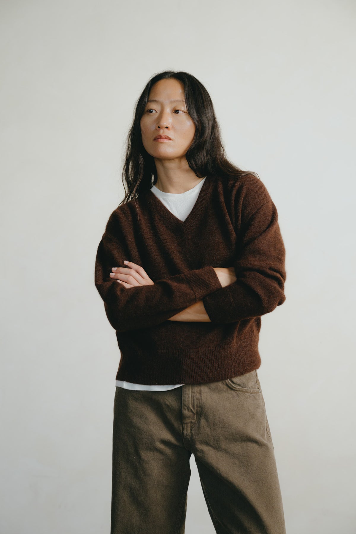 A person with long dark hair stands against a plain light gray background wearing a brown V-neck sweater over a white undershirt and loose brown pants, with arms crossed.