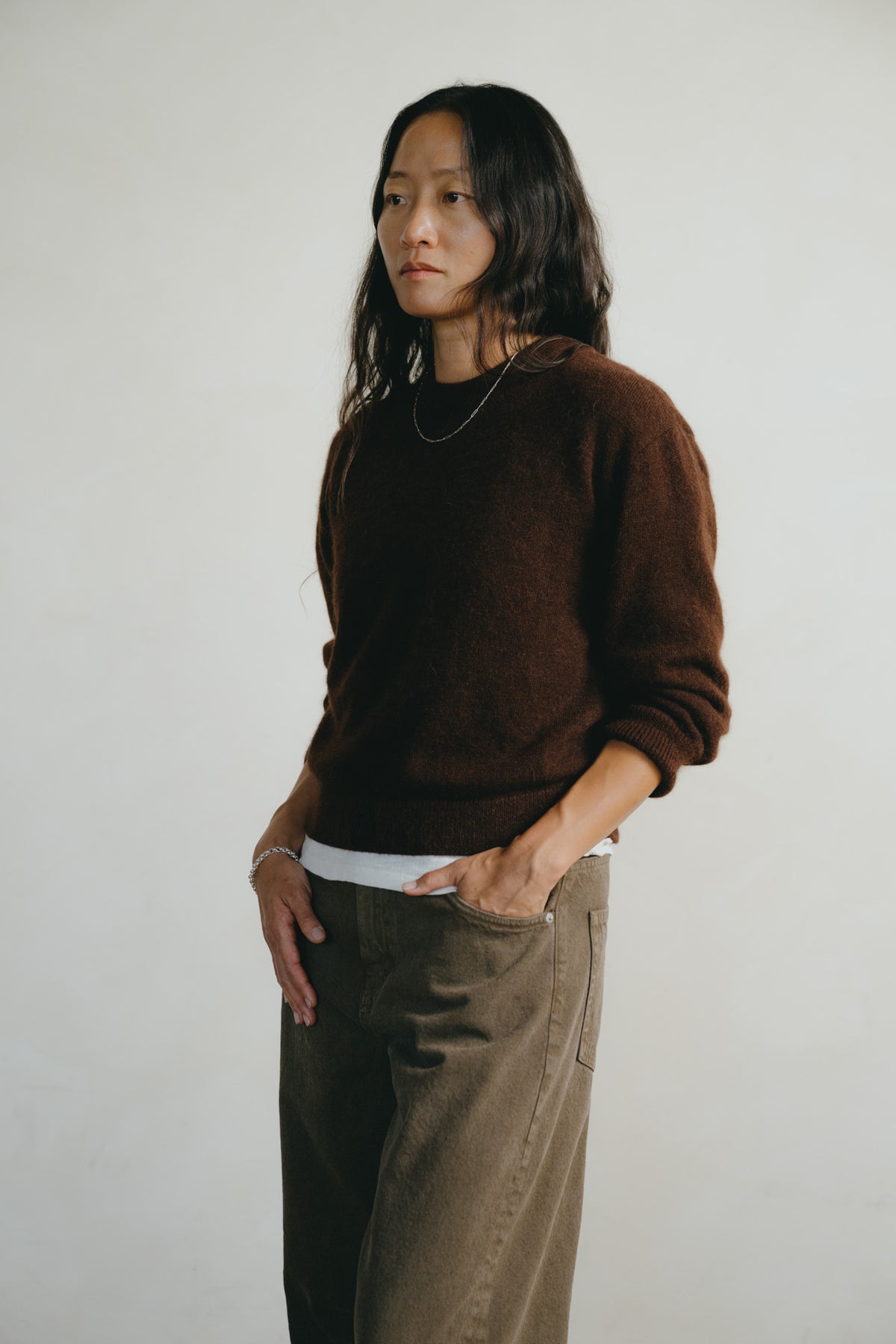 A model wearing a dark brown knit crewneck sweater over a white undershirt and brown wide-leg pants, standing against a plain off-white wall.