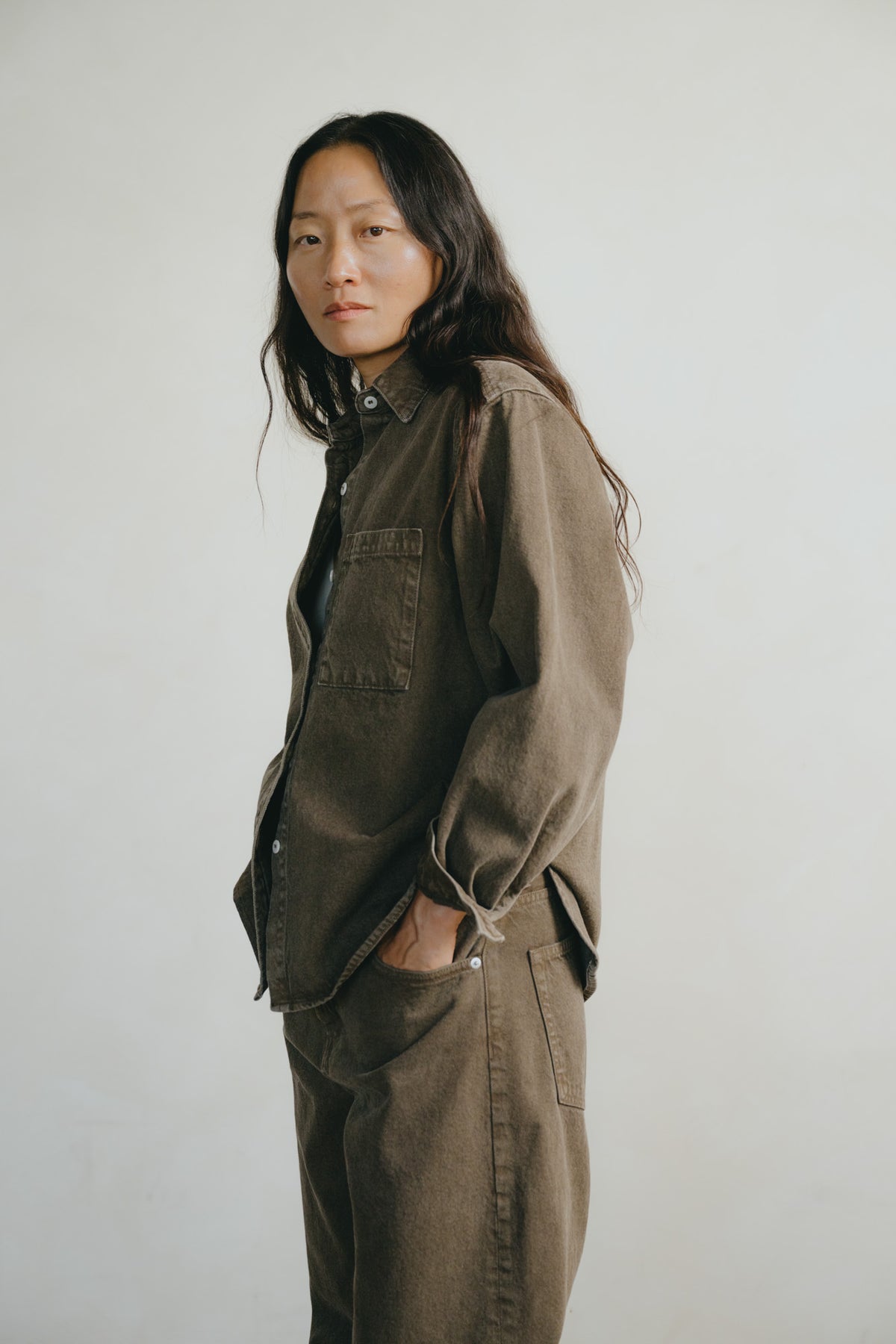 A woman with long dark hair stands in profile wearing an oversized brown corduroy utility shirt and matching brown corduroy pants against a plain light gray background.