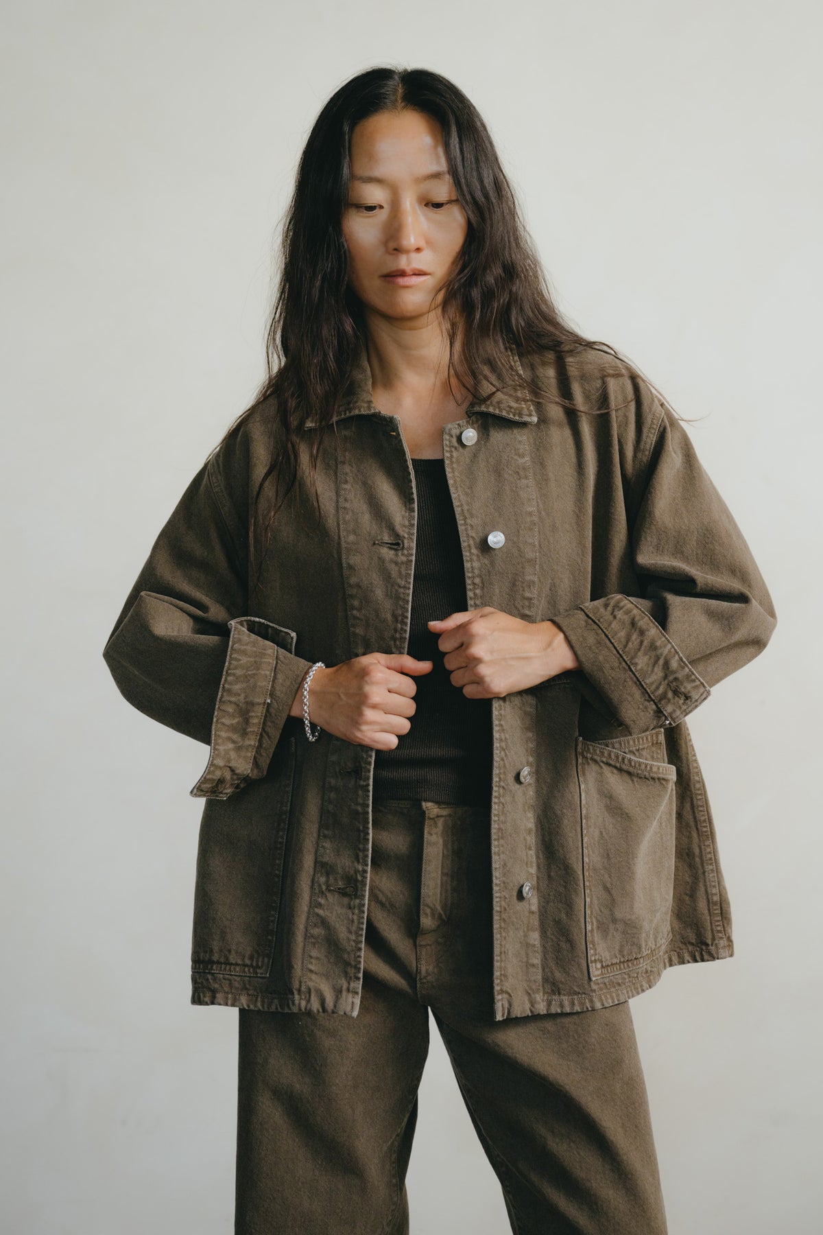 A woman wearing an oversized brown utility jacket over a black top and matching brown pants, standing against a plain light gray background.