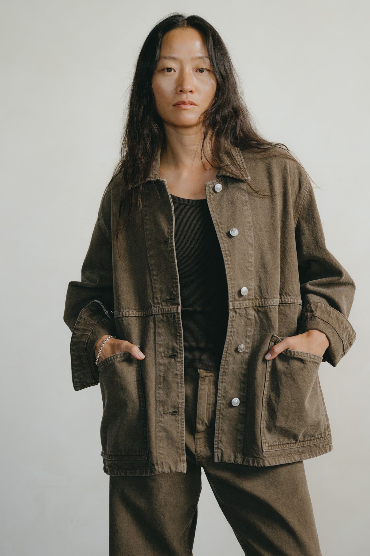A woman wearing an oversized brown utility jacket over a black top and matching brown pants, standing against a plain light gray background.