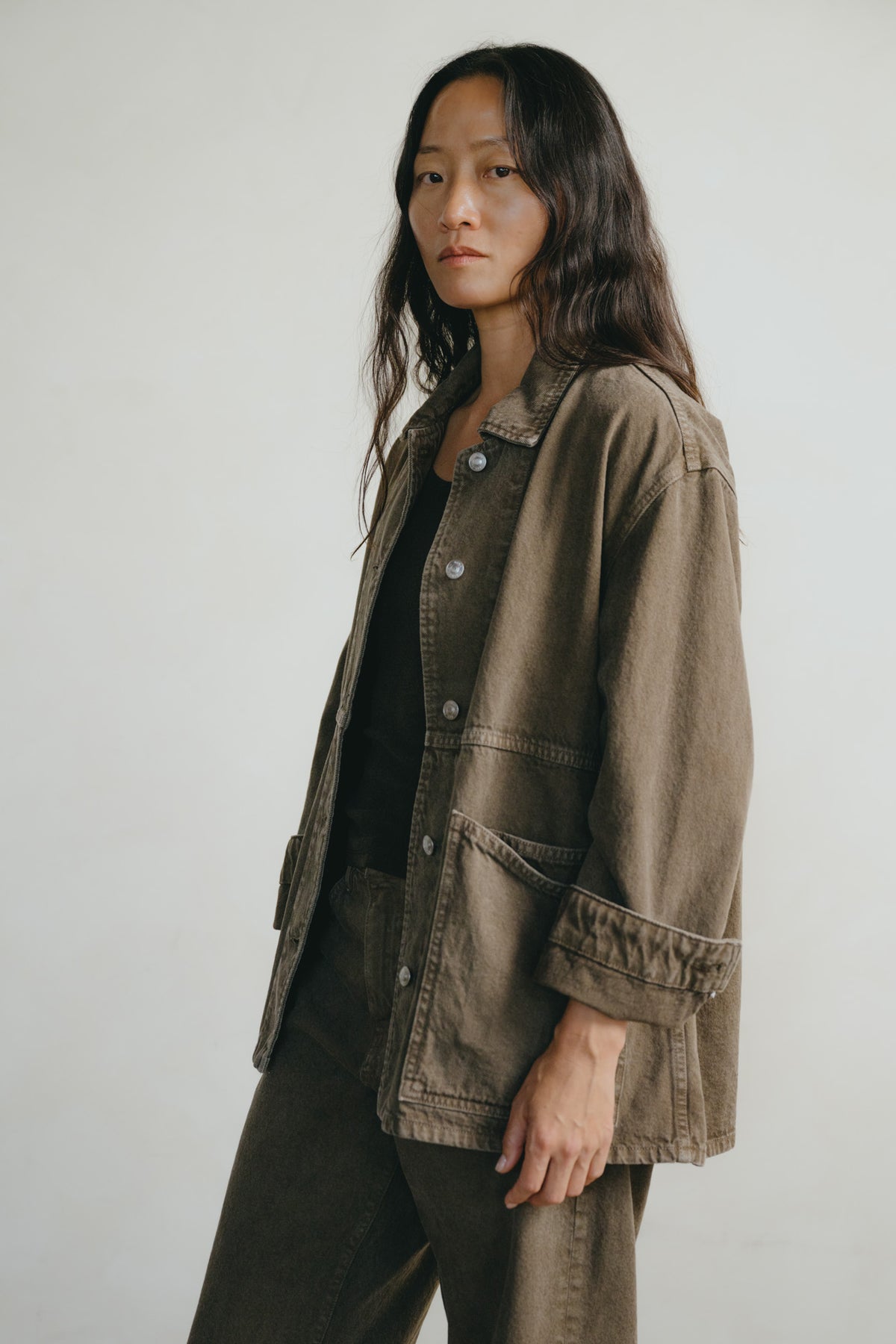 A woman wearing a dark brown utility jacket over a black top and matching dark brown pants, standing against a plain light gray studio background.