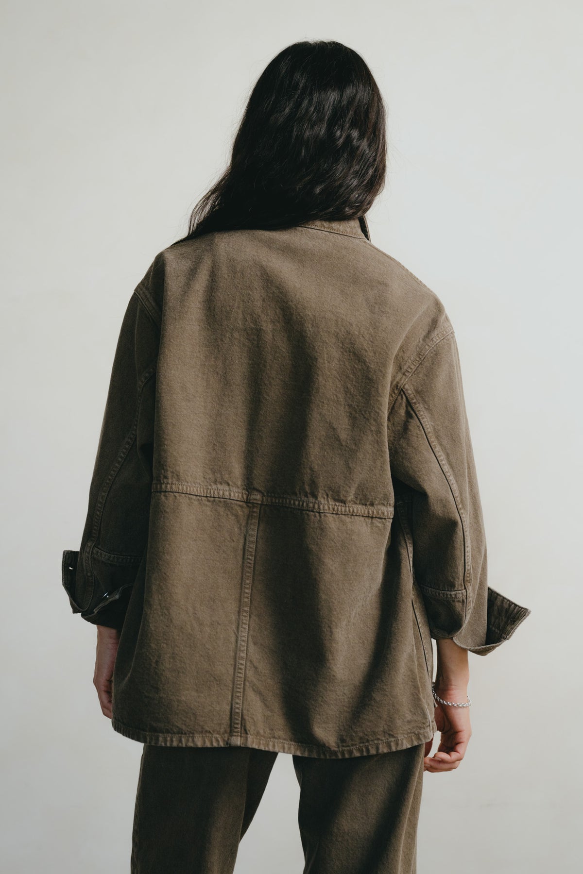 A person shown from the back wearing an oversized brown utility jacket with three-quarter sleeves and matching brown pants, photographed against a plain light gray studio background.