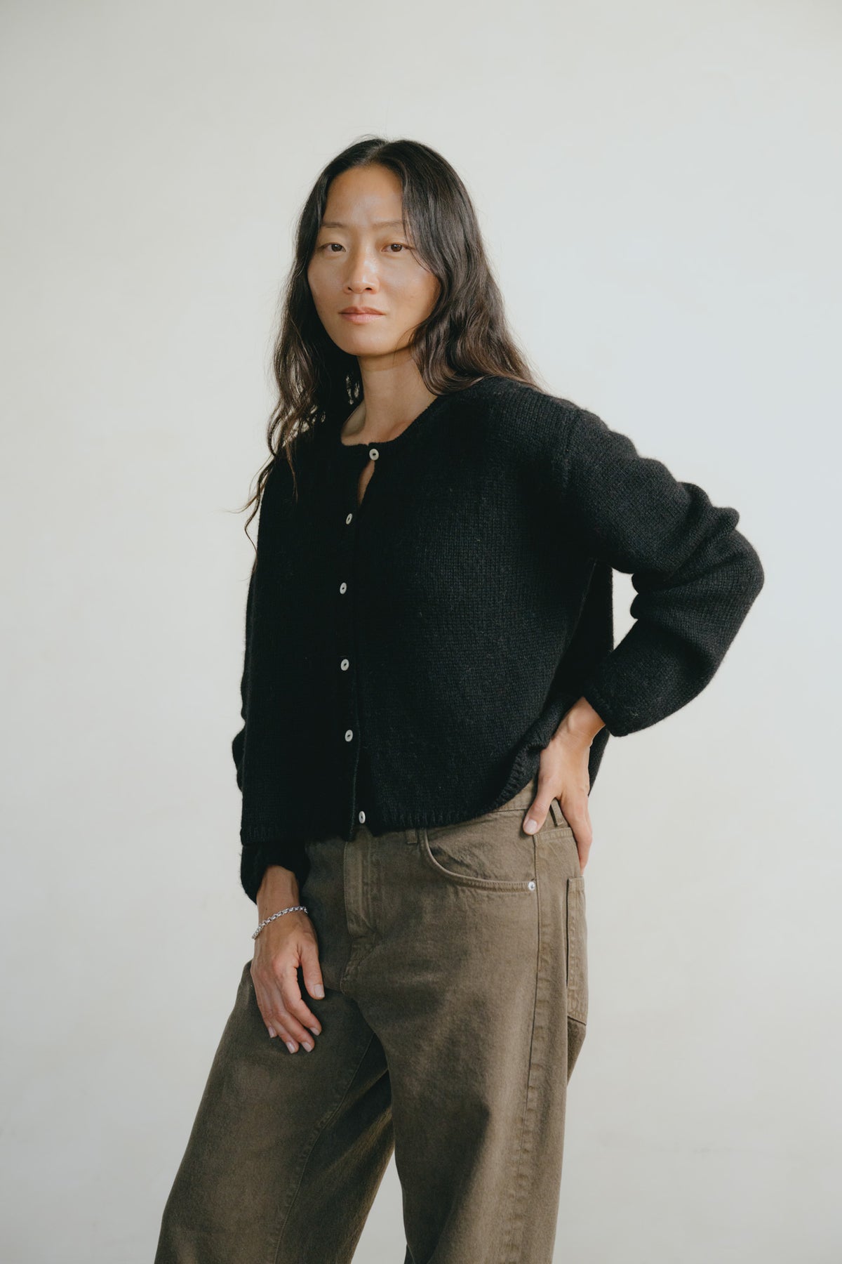 A woman modeling a black button-up knit cardigan and brown wide-leg pants against a plain light gray studio background.