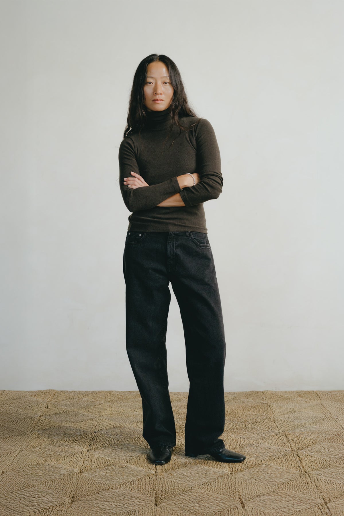 A woman stands with arms crossed wearing a dark brown ribbed turtleneck sweater and black wide-leg jeans against a plain off-white wall and textured tan floor.