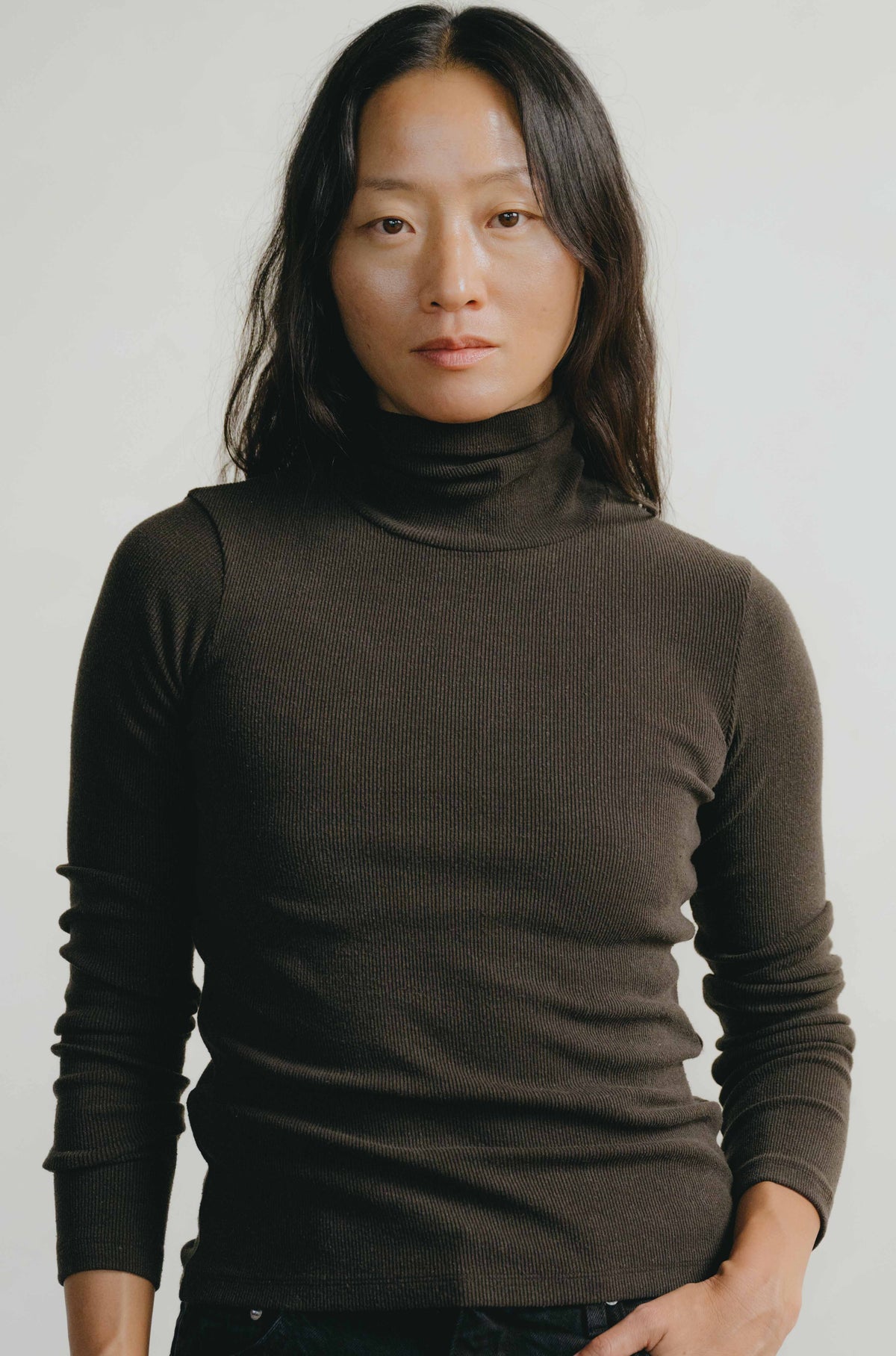A woman wearing a dark brown ribbed turtleneck sweater, photographed from the chest up against a plain light gray background.