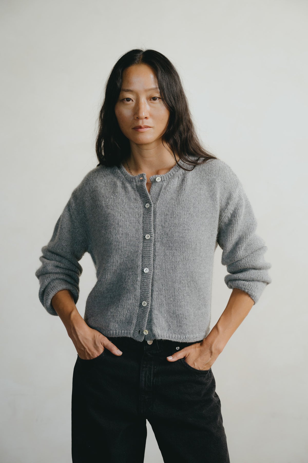 A woman wearing a gray knit button-front cardigan and black jeans, standing against a plain light gray background with her hands in her pockets.