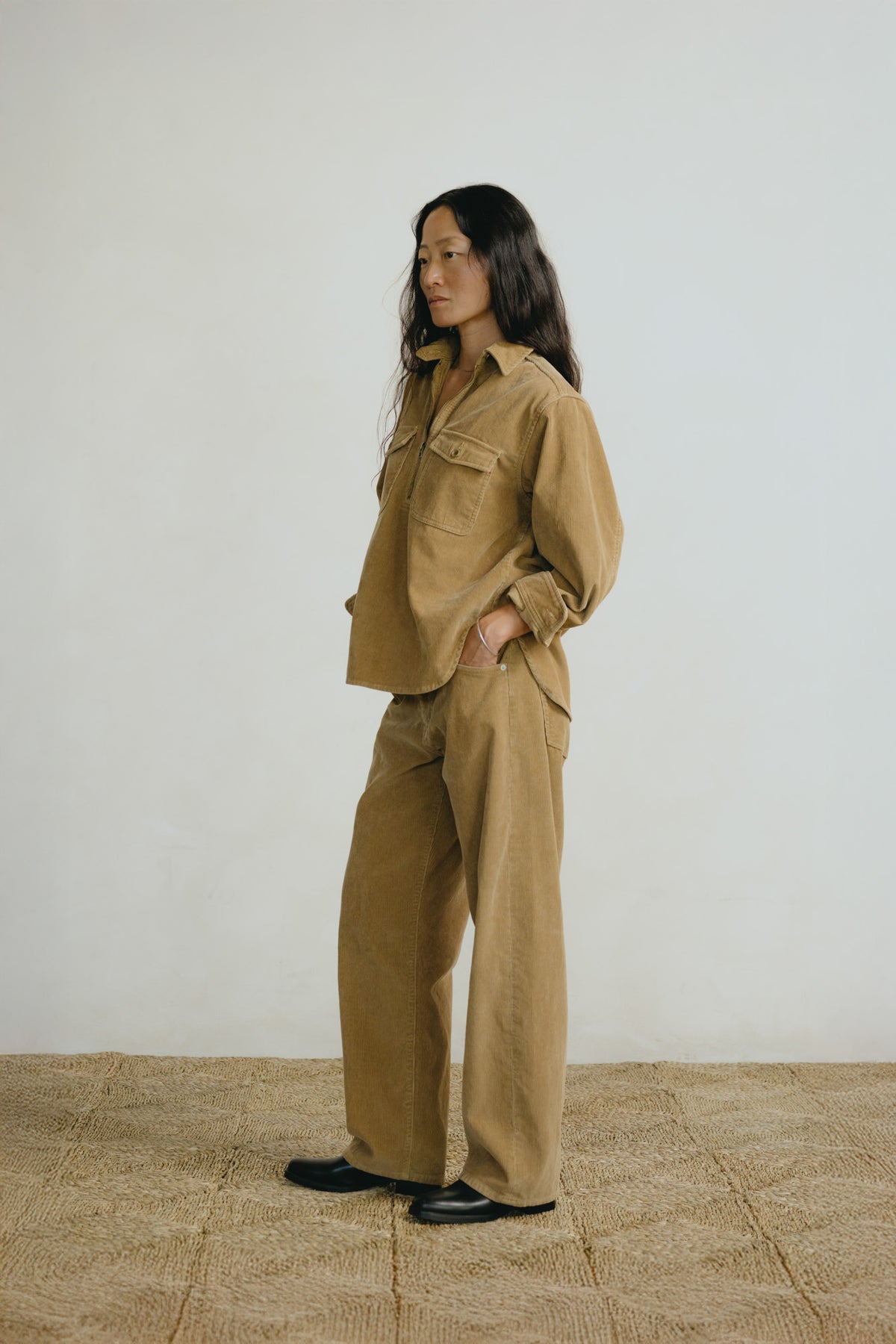 A woman stands in profile wearing a matching tan utility jacket and wide-leg trousers set, paired with black shoes, against a plain off-white wall and textured tan floor.
