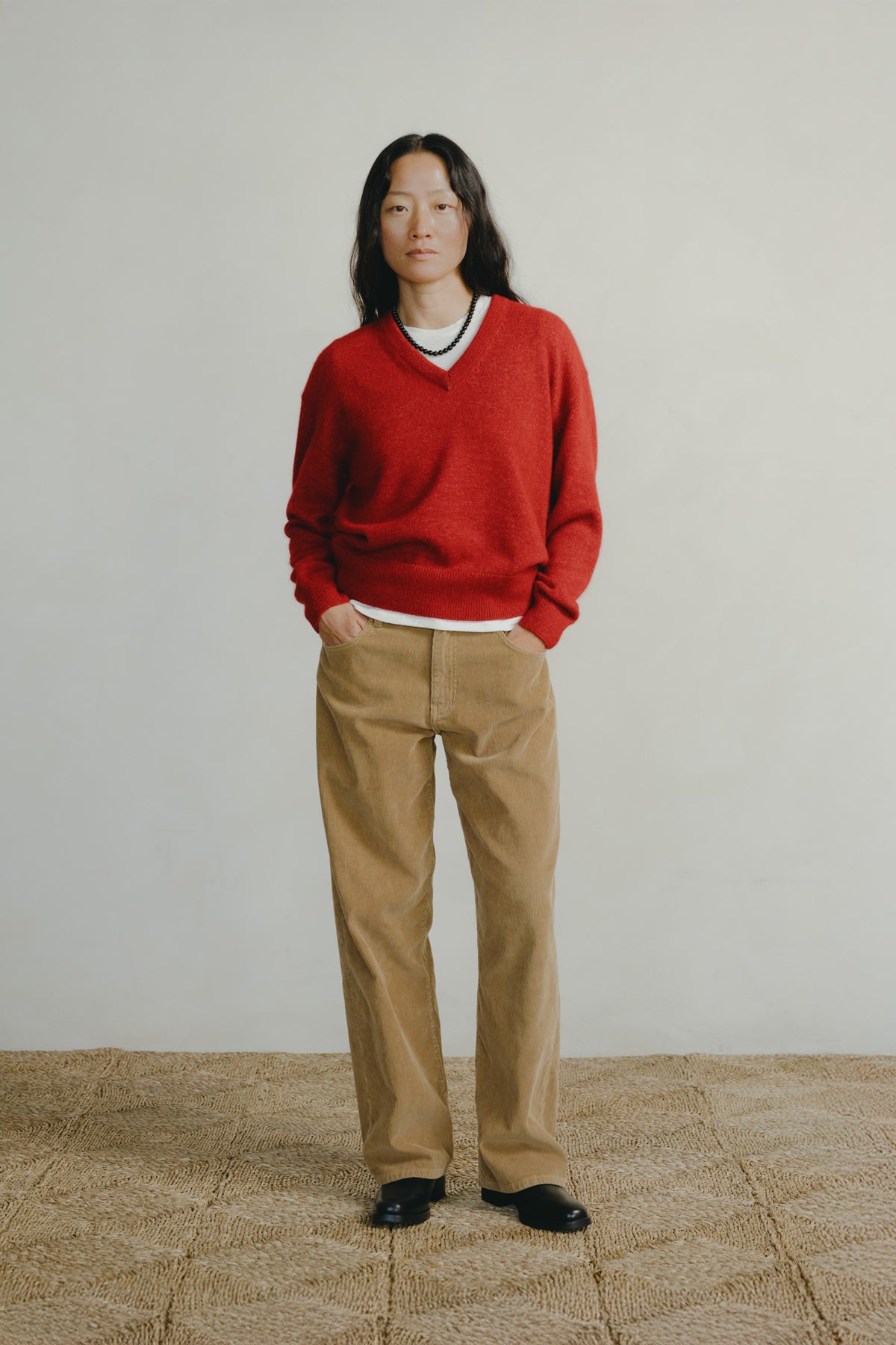 A model stands against a plain light gray wall wearing a bright red V-neck knit sweater over a white collared shirt, paired with tan trousers and black shoes.