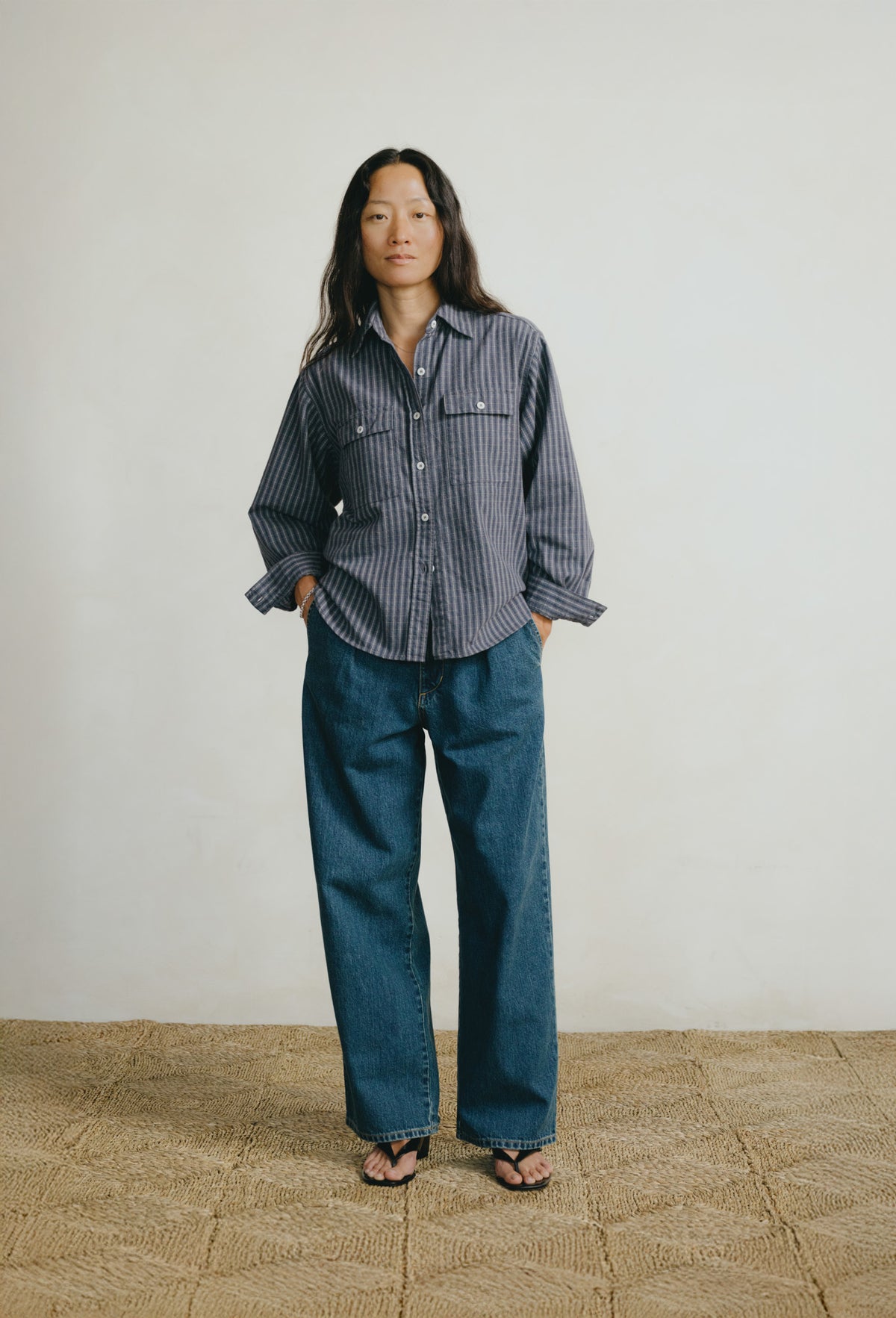 A model stands against a plain off-white wall wearing a dark blue and gray vertically striped button-down shirt, wide-leg blue jeans, and black sandals.