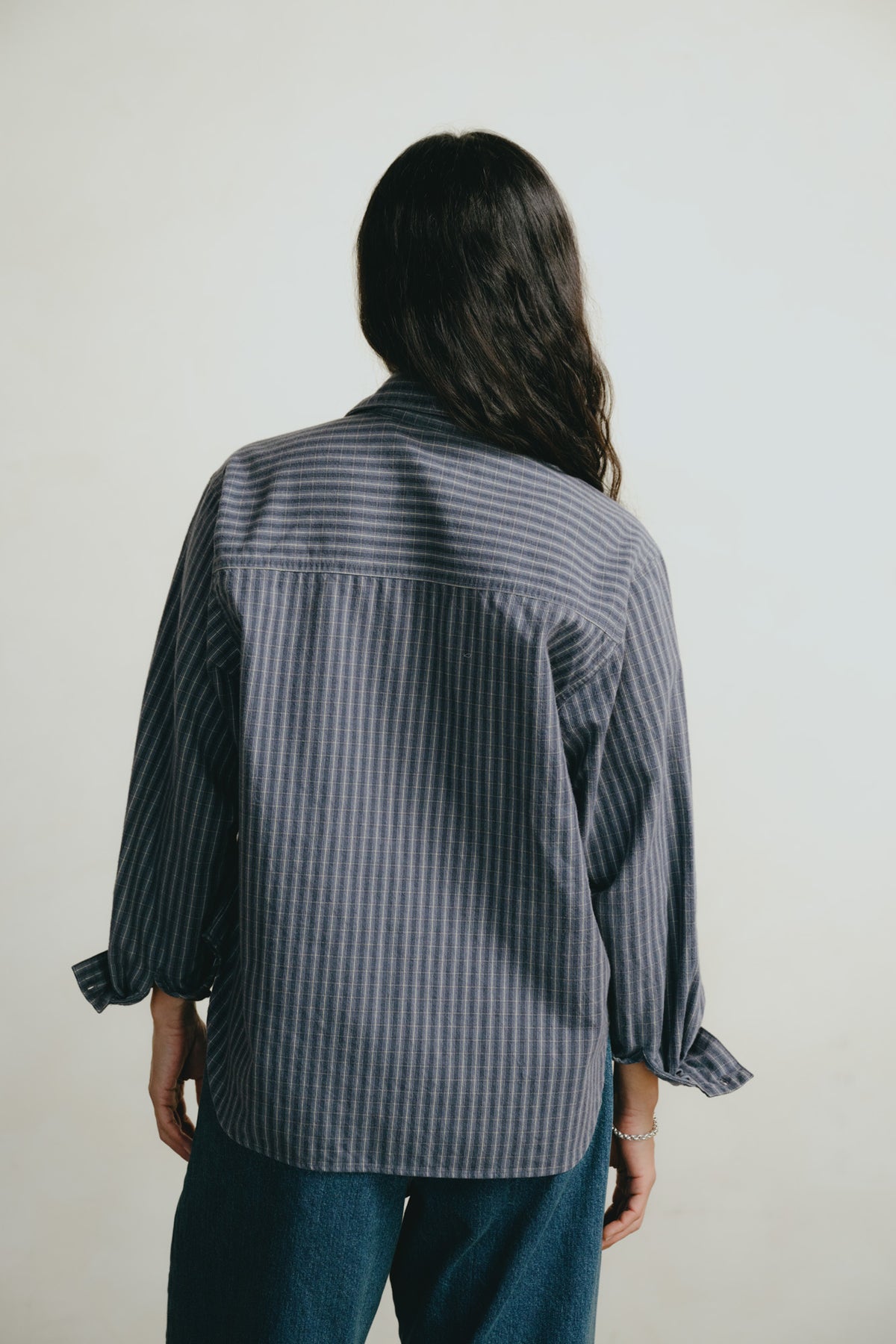 A person viewed from behind wearing a dark blue and gray vertically striped button-up shirt with rolled-up sleeves and dark blue jeans, set against a plain off-white background.