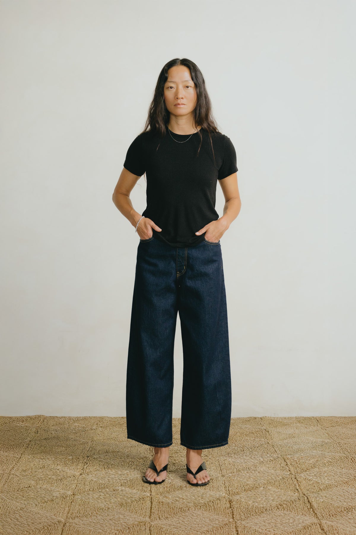 A woman stands against a plain off-white wall wearing a black short-sleeve crewneck t-shirt, dark wide-leg denim jeans, and black sandals.