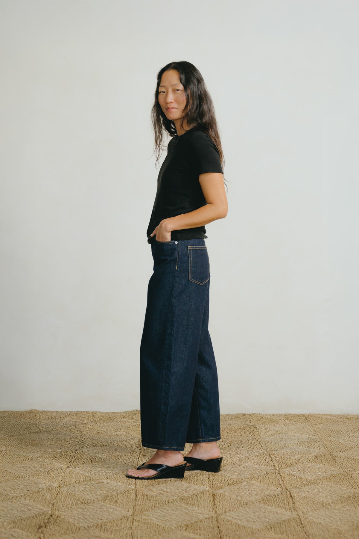 A woman stands in profile wearing dark wide-leg denim jeans, a black short-sleeve top, and black sandals against a plain off-white wall and woven tan floor.