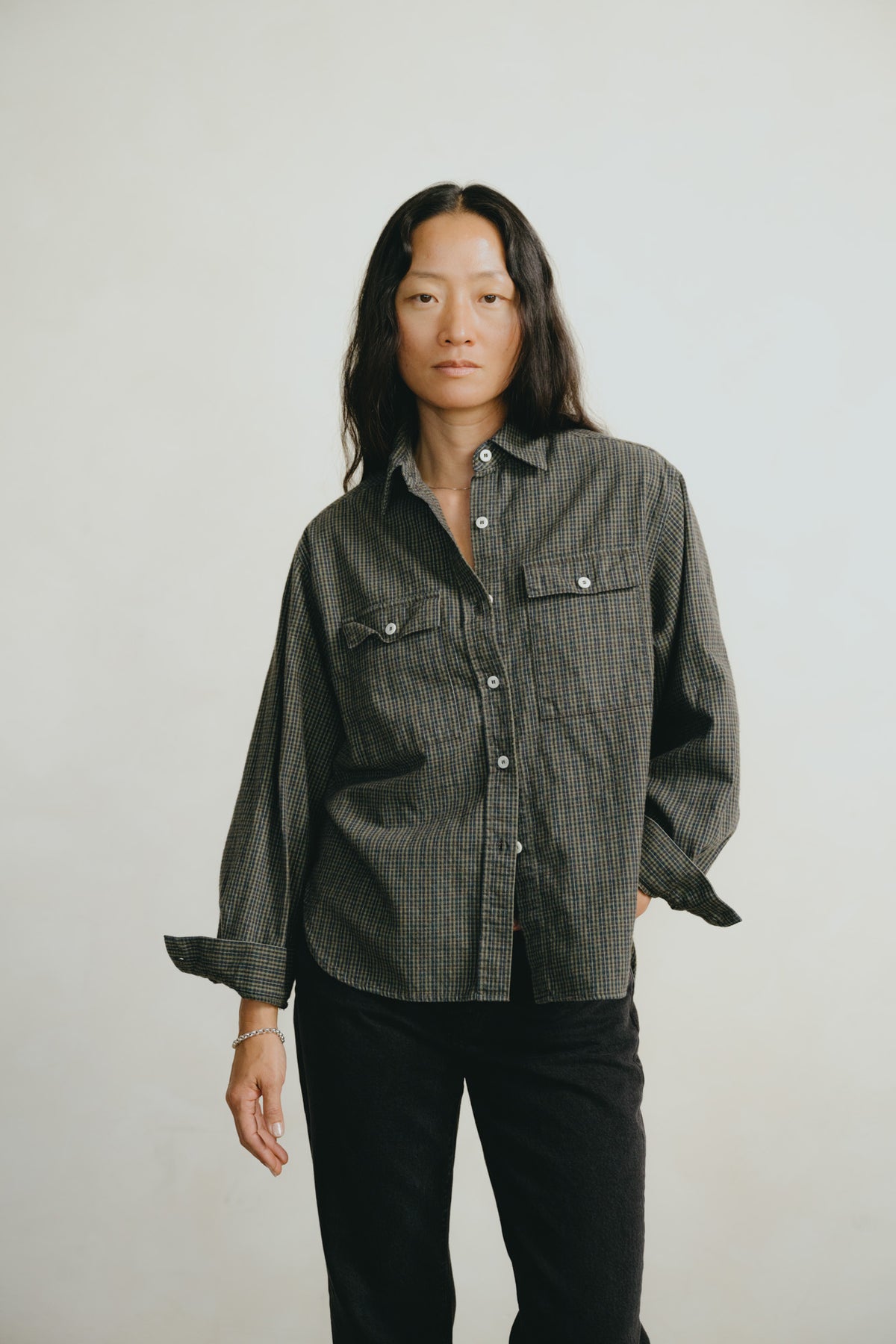 A model stands against a light gray studio background wearing a dark gray and black plaid button-down shirt with black pants.