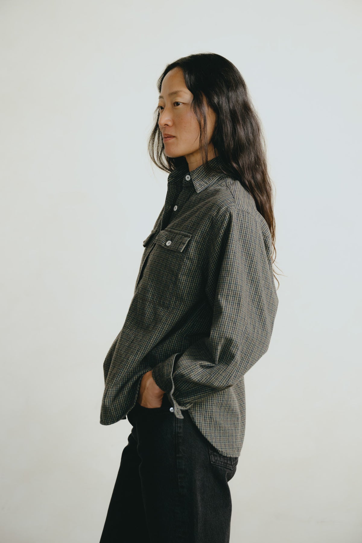 A woman with long dark hair is shown in profile wearing a dark gray and black checkered button-up shirt and black pants, standing against a plain light gray studio background.