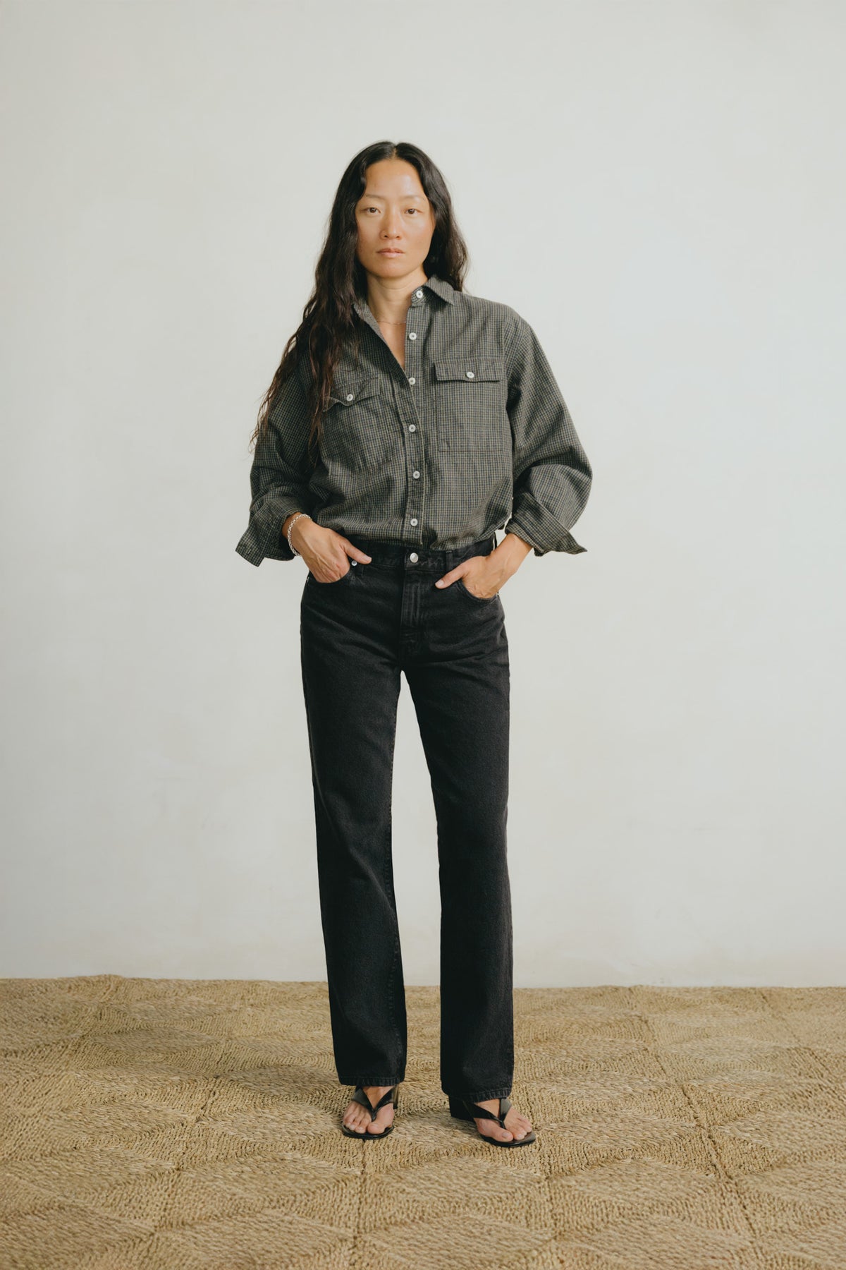 A woman stands against a plain light gray wall wearing a dark gray button-up shirt with chest pockets, black jeans, and black sandals, with her hands in her pockets.