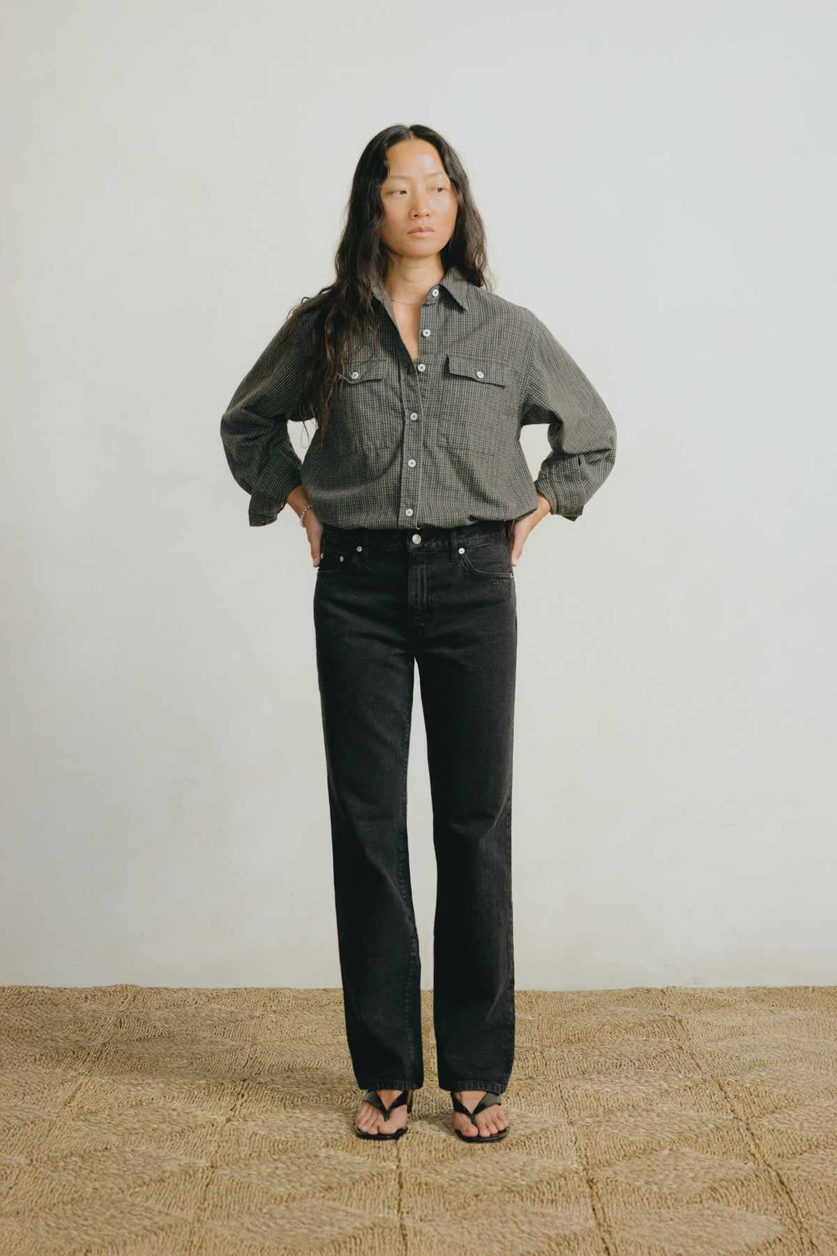 A model stands against a light gray wall and textured tan floor, wearing a dark gray and black striped button-up shirt, black jeans, and black sandals.