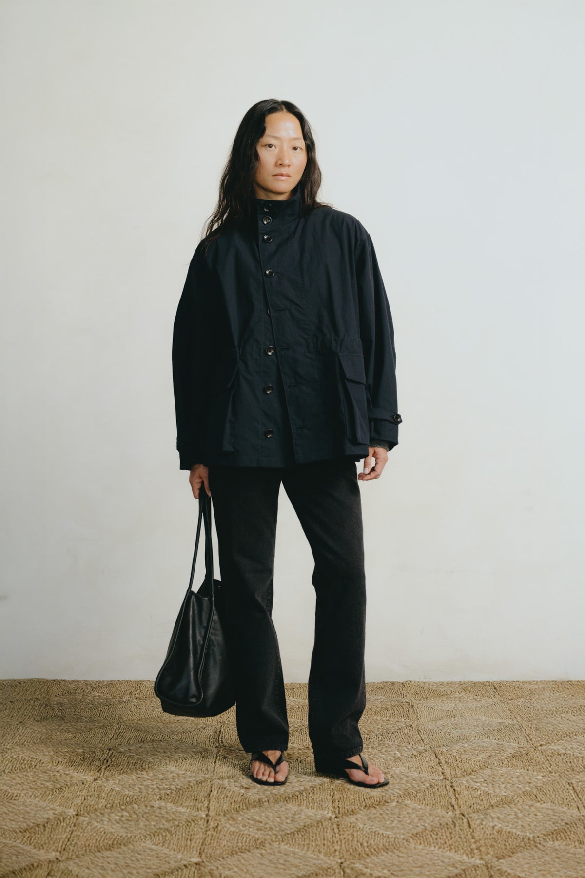 A model stands against a plain off-white wall wearing an oversized black jacket, black wide-leg pants, and black sandals, while holding a black tote bag.