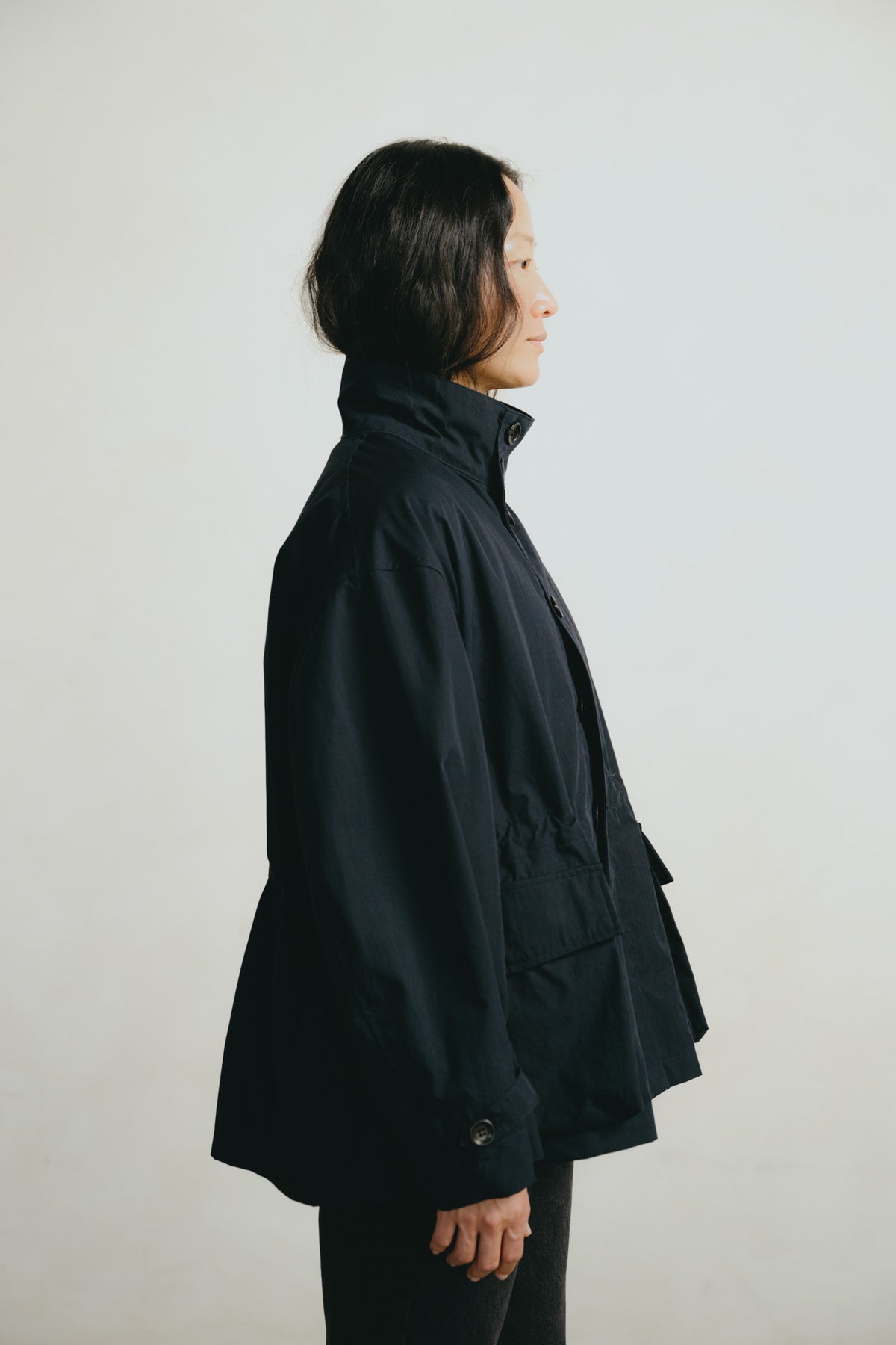A woman shown in profile wearing a dark navy or black lightweight jacket against a plain light gray studio background.
