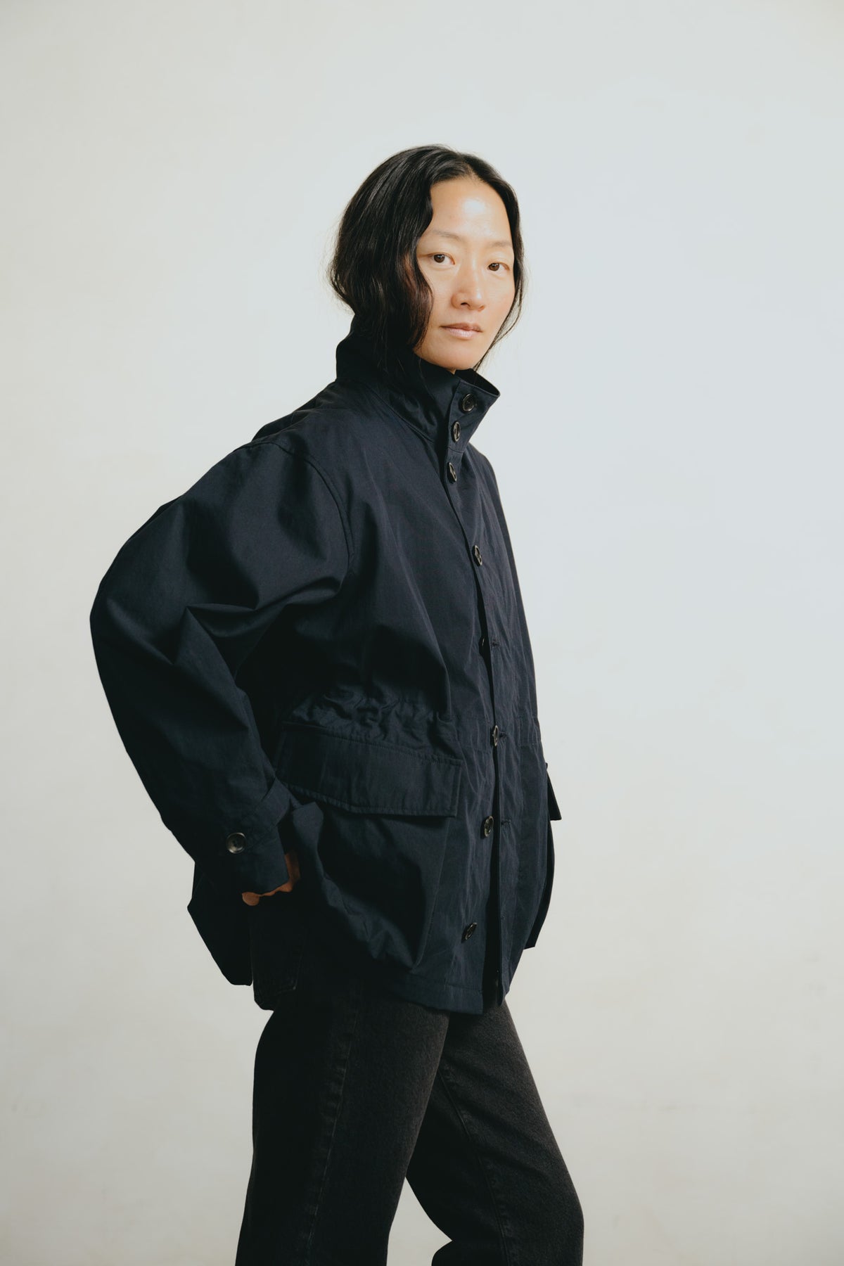 A model wearing a dark navy blue utility jacket with a high collar and front pockets, paired with black pants, standing against a plain light gray background.