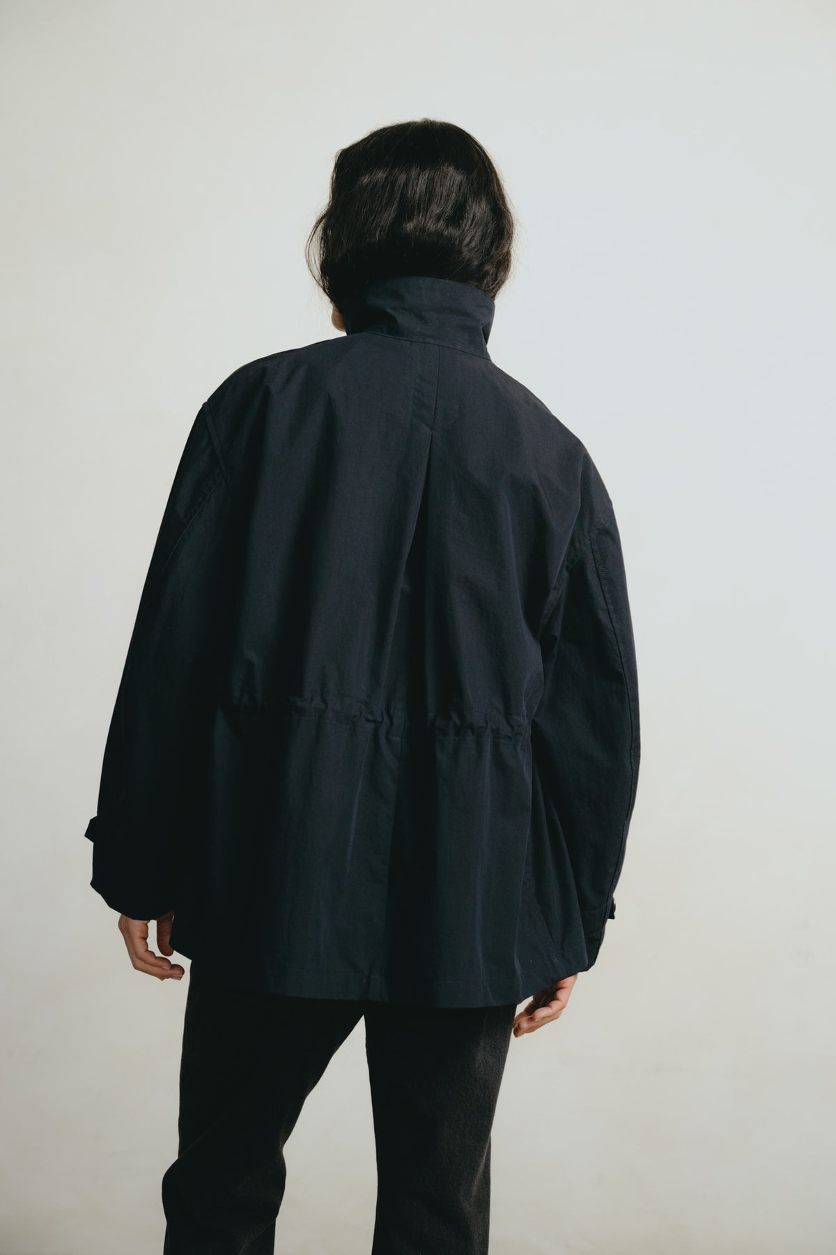 A person viewed from behind wearing a dark navy blue jacket with a high collar and a gathered waist, paired with black pants, against a plain off-white background.