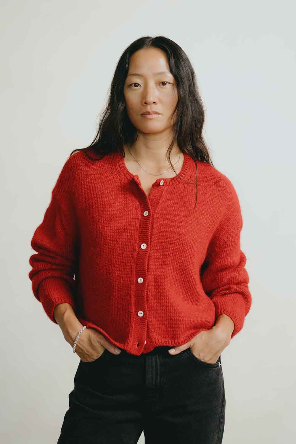 A woman wearing a textured red button-up cardigan sweater and black jeans, standing against a plain light gray background.
