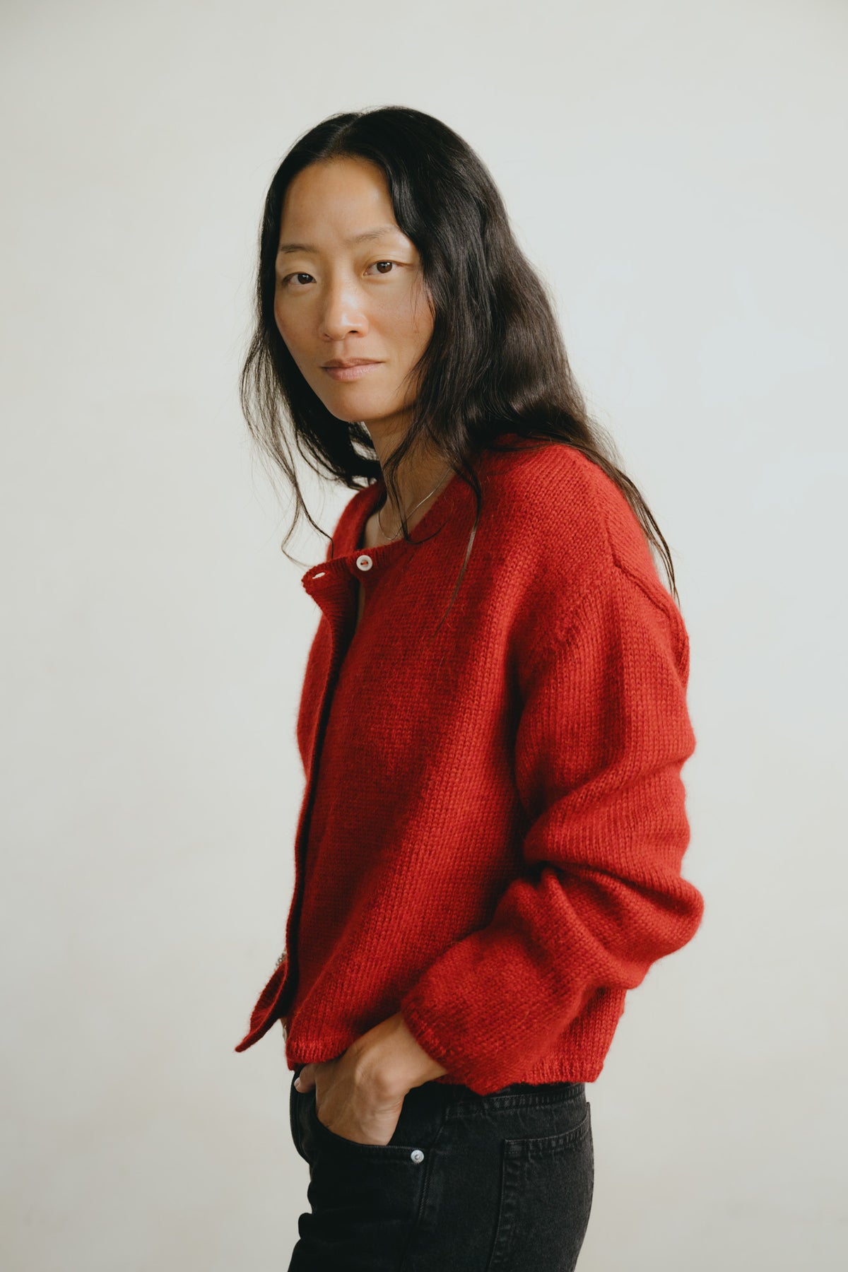 A woman with long dark hair wearing a textured red knit cardigan and black jeans, standing against a plain light gray background.
