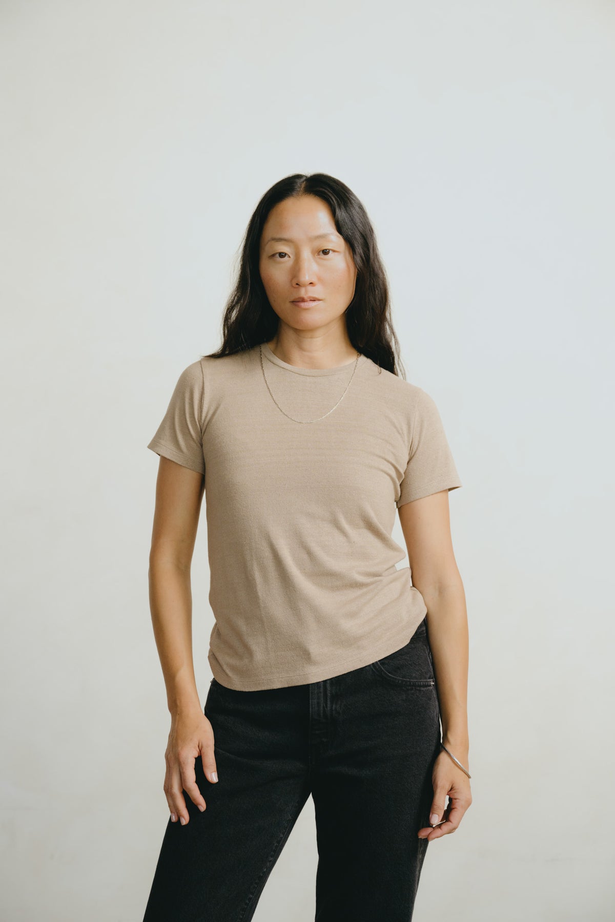A woman with long dark hair stands against a light gray studio background wearing a beige short-sleeve crewneck t-shirt and black jeans.