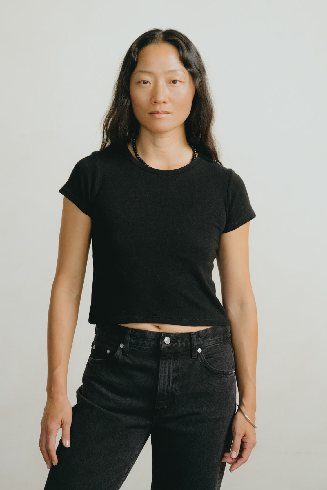 A woman modeling a black short-sleeve cropped t-shirt and black jeans against a plain light gray studio background.