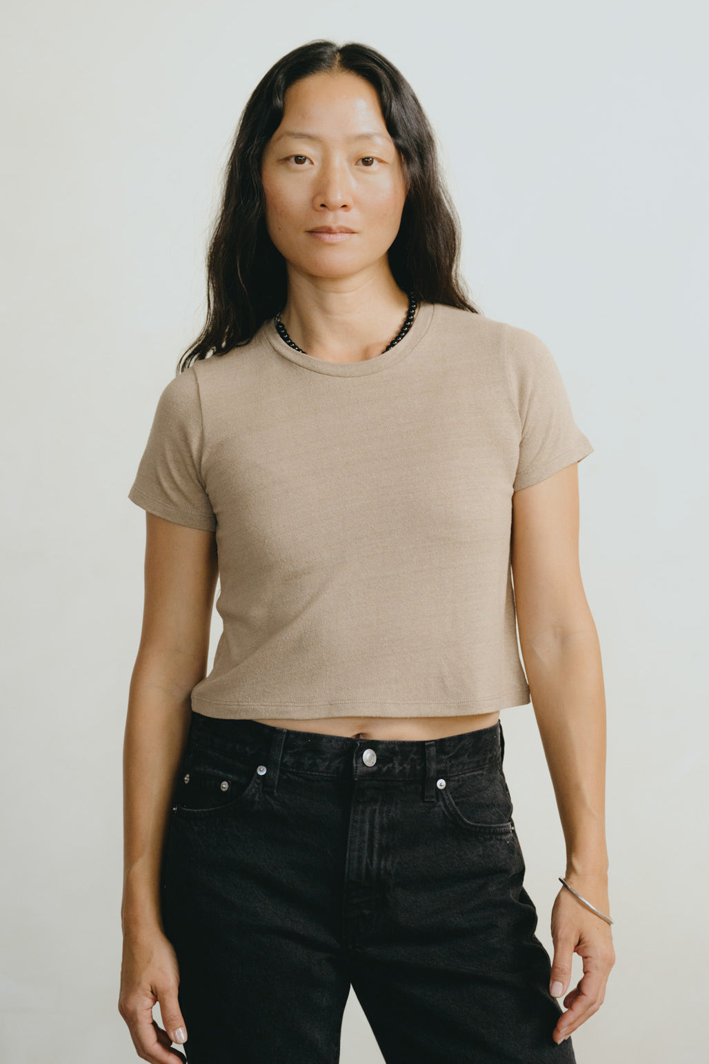 A woman modeling a cropped short-sleeve crewneck t-shirt in a light beige color, paired with black jeans, against a plain light gray background.