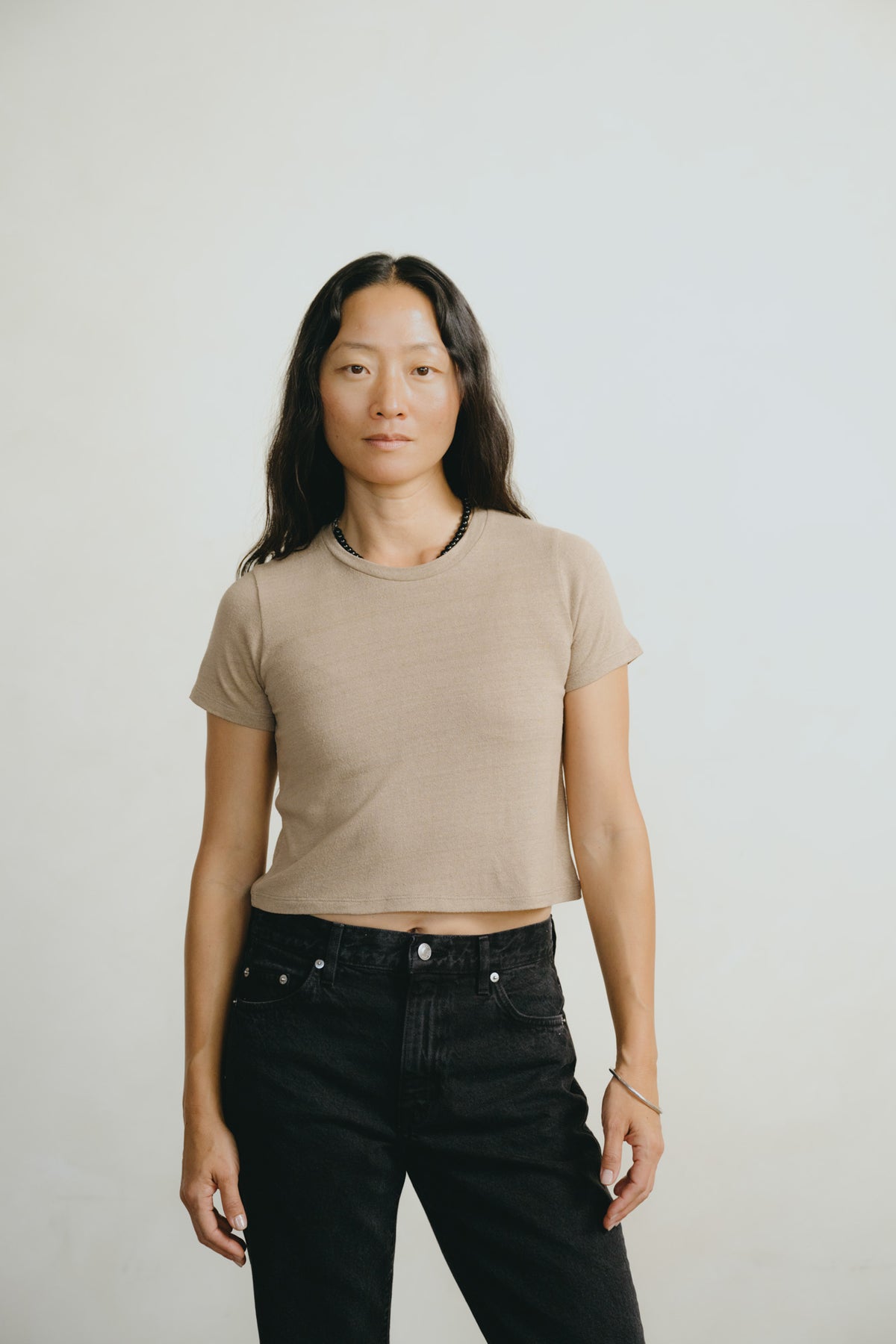A woman modeling a beige short-sleeve cropped t-shirt and black jeans against a plain light gray background.