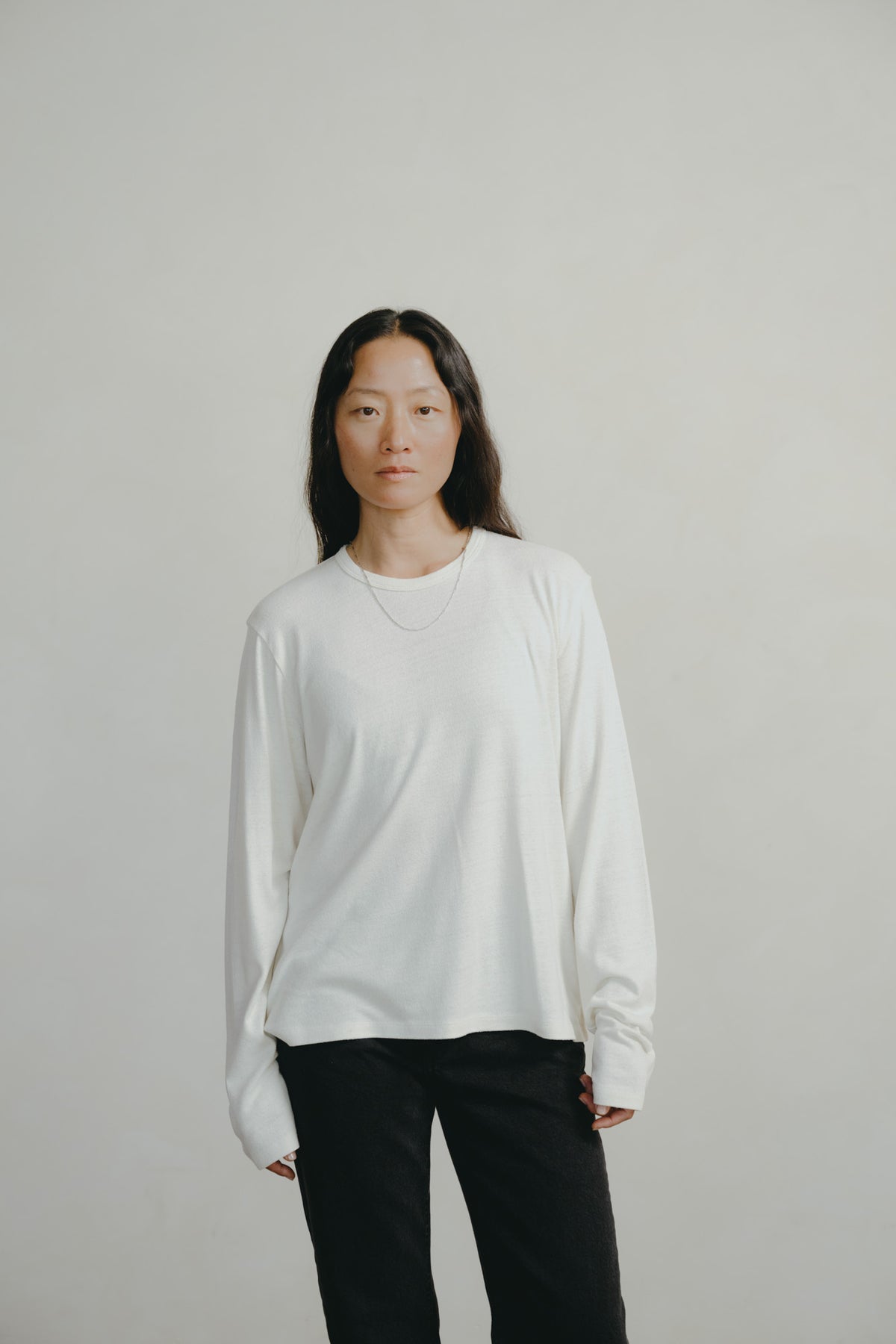 A woman modeling a white long-sleeve crewneck shirt with black pants, standing against a plain light gray studio background.