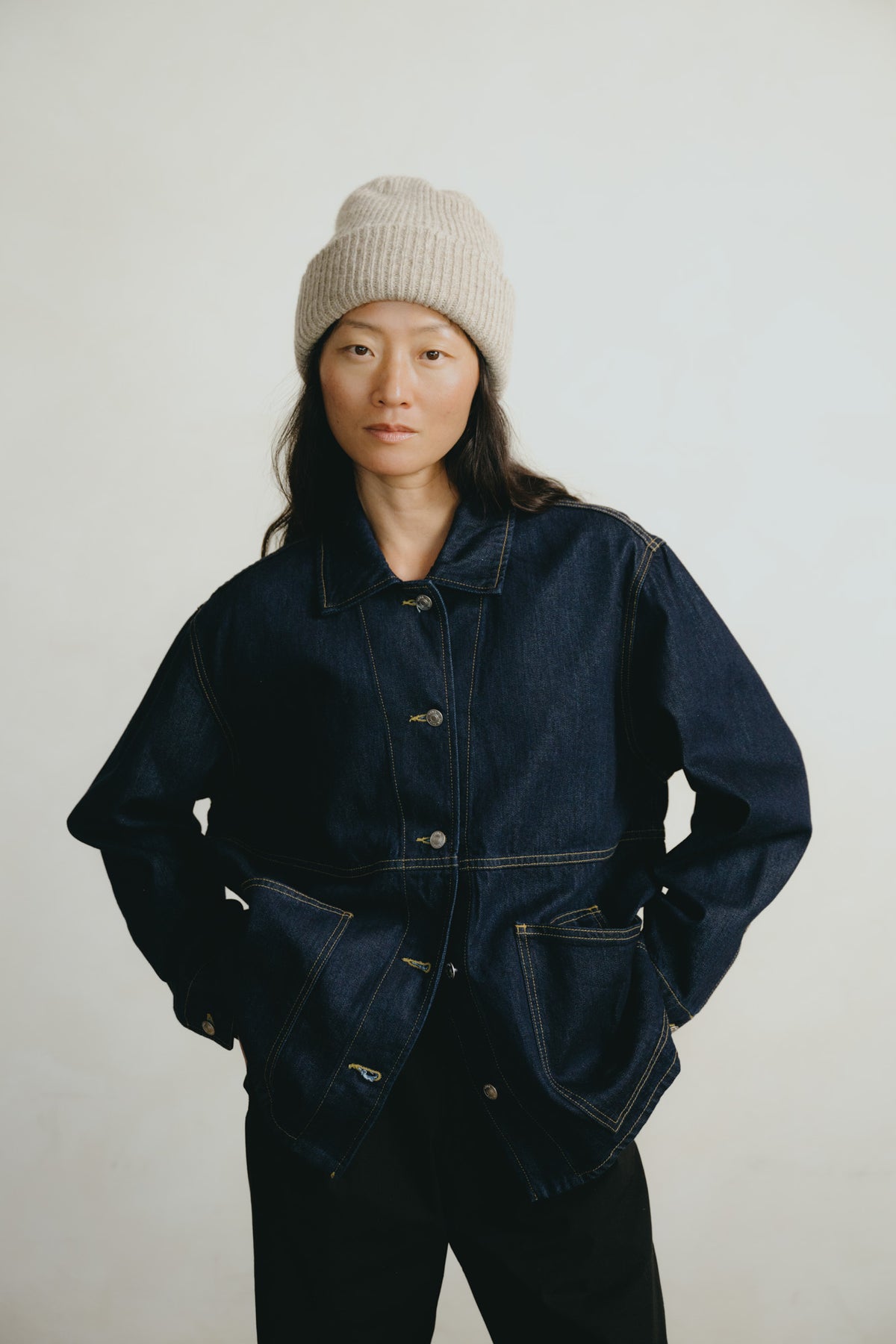 A model wearing a dark indigo denim utility jacket with contrast yellow stitching, a light beige knit beanie, and black pants, standing against a plain light gray background.
