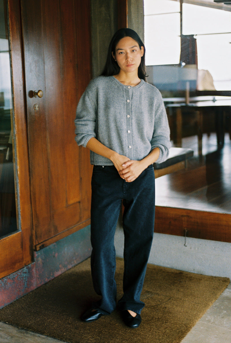 A woman stands indoors wearing a light gray button-front cardigan, dark wide-leg jeans, and black flats, positioned in front of a rustic wooden door and window.