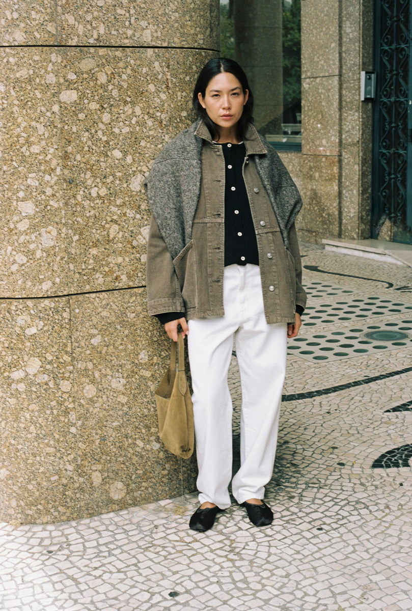 A model stands outdoors against a textured stone wall, wearing a layered outfit consisting of a brown tweed jacket over a black top, white trousers, and black loafers, while holding a tan tote bag.