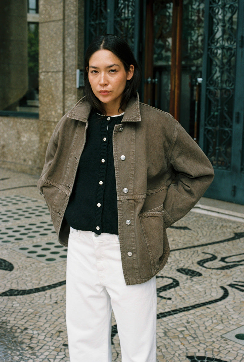 A woman wearing a brown corduroy utility jacket over a black button-up top and white trousers, standing outdoors on a patterned stone pavement in front of a building entrance.