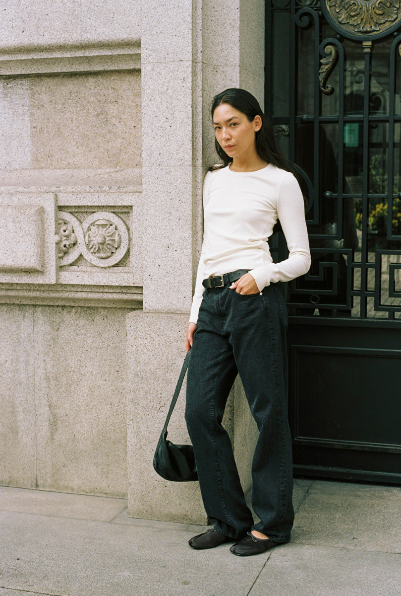 A woman standing in a doorway with a long sleeve tight white shirt and black jeans.