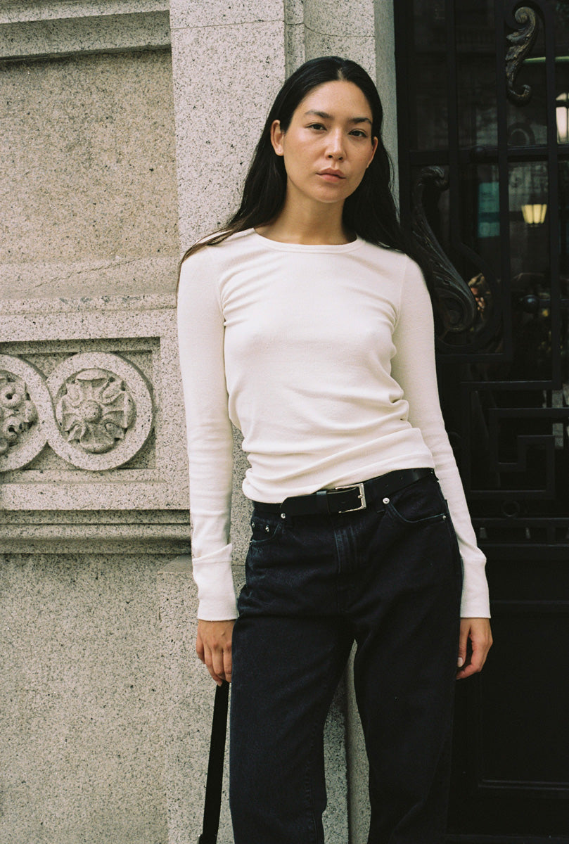 A woman standing in a doorway with a long sleeve tight white shirt and black jeans.