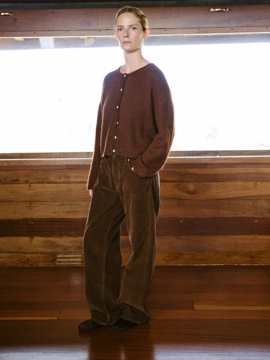 A woman stands in a rustic indoor setting with wood paneling and a wooden floor, wearing a brown button-front knit cardigan and matching brown corduroy pants.