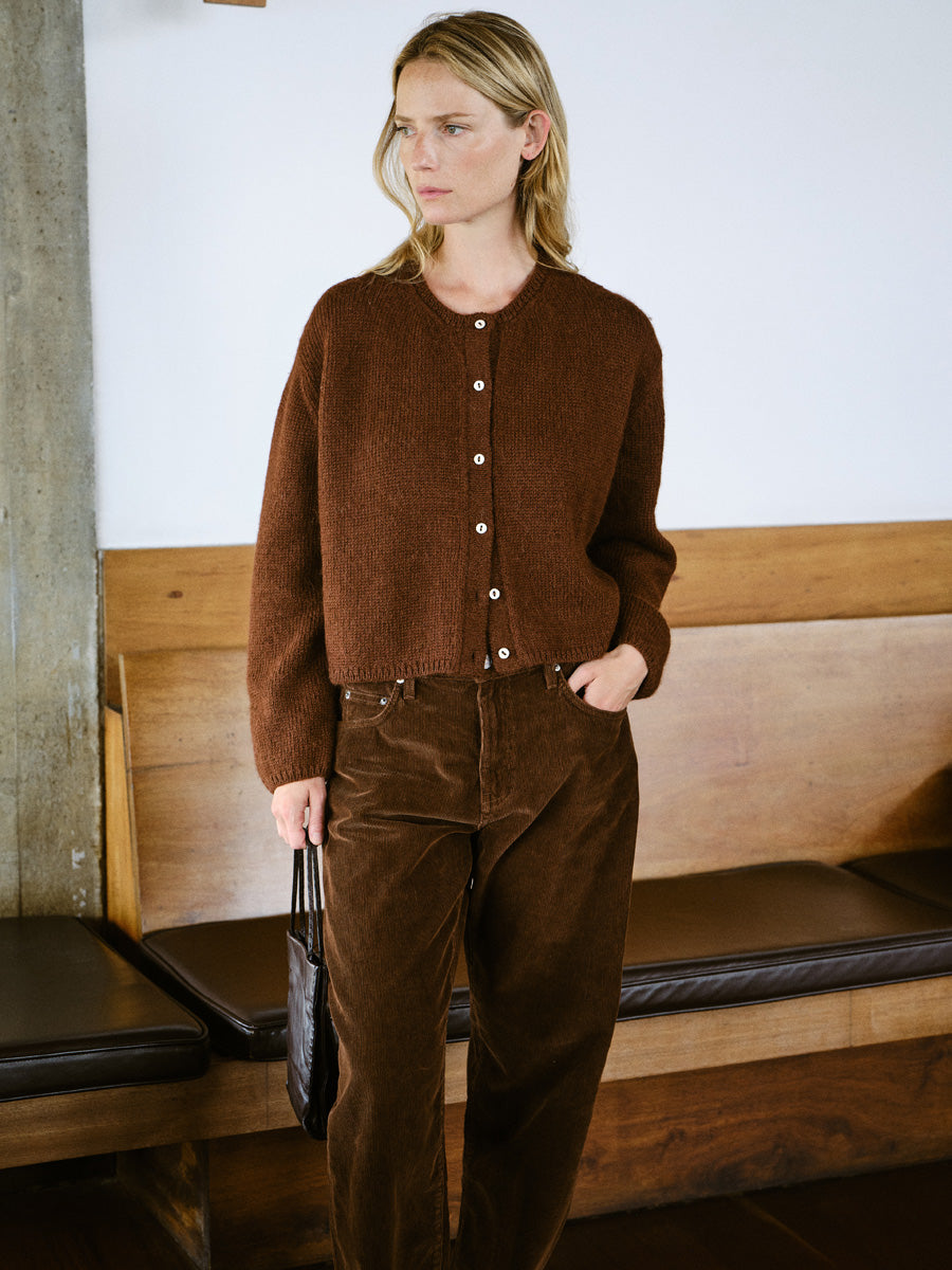 A woman stands indoors wearing a brown button-front knit cardigan and matching brown corduroy pants, holding a black handbag. The setting includes a wooden bench and a light wall.