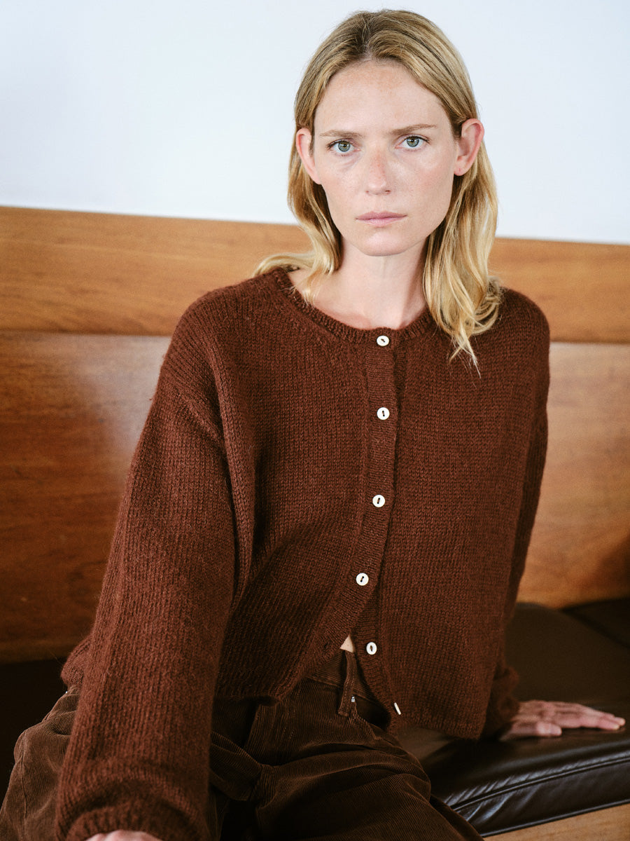 A woman with blonde hair sits on a wooden bench wearing a brown textured knit button-up cardigan and brown pants.