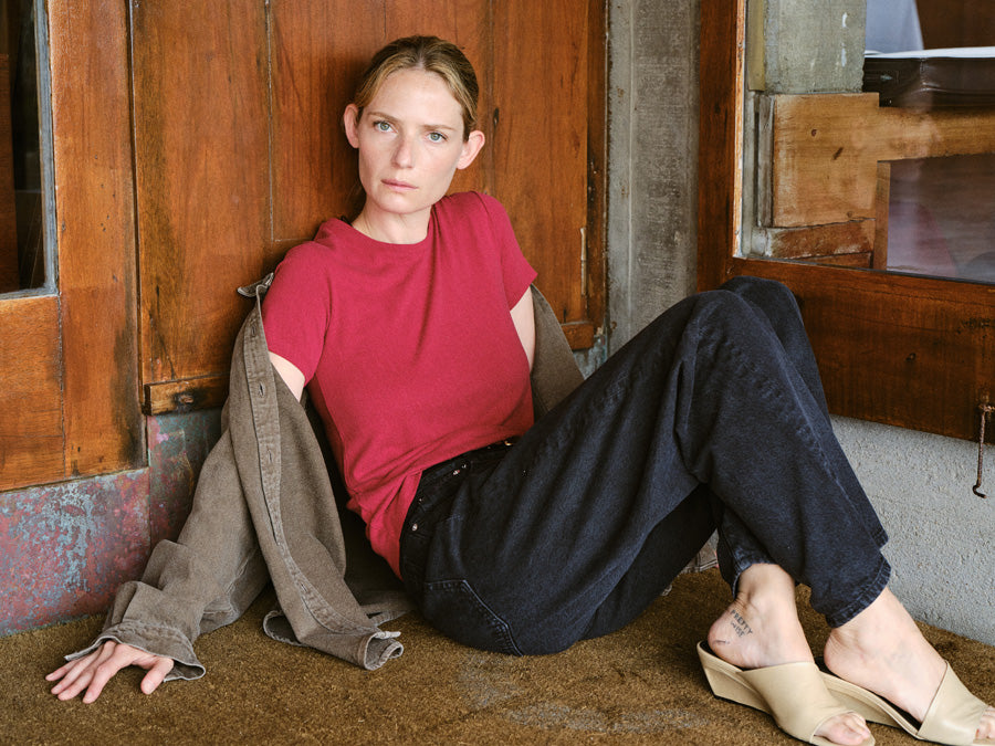 A woman sitting against a wood doorway in a red crewneck tshirt.