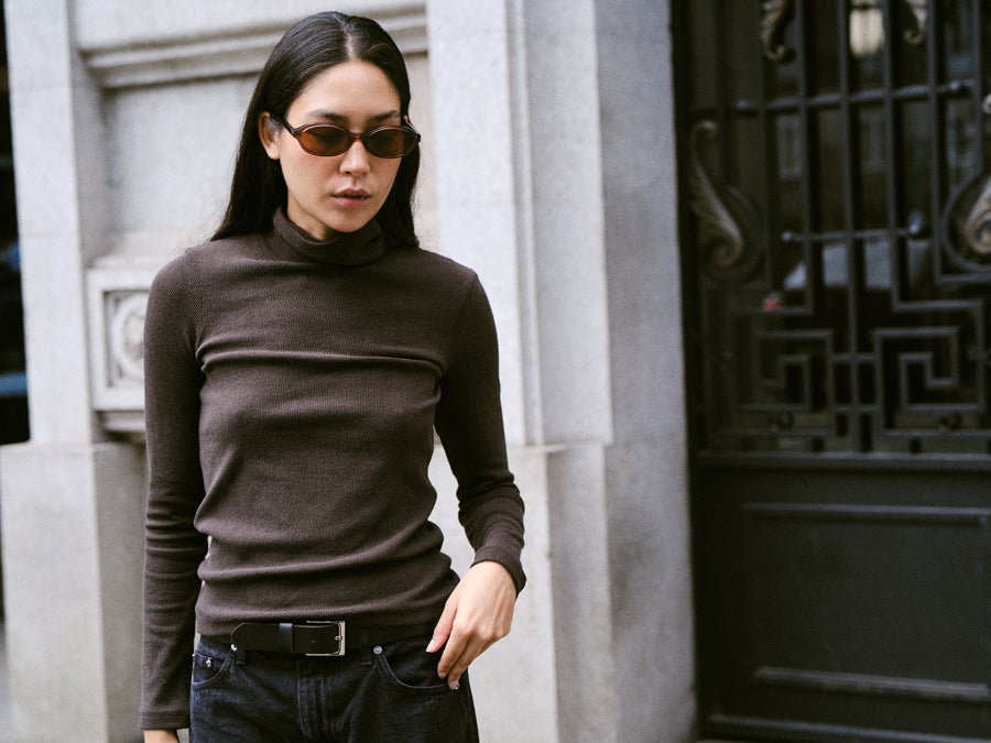 A woman standing on the street with sunglasses and wearing a long-sleeved dark brown turtleneck and black jeans.